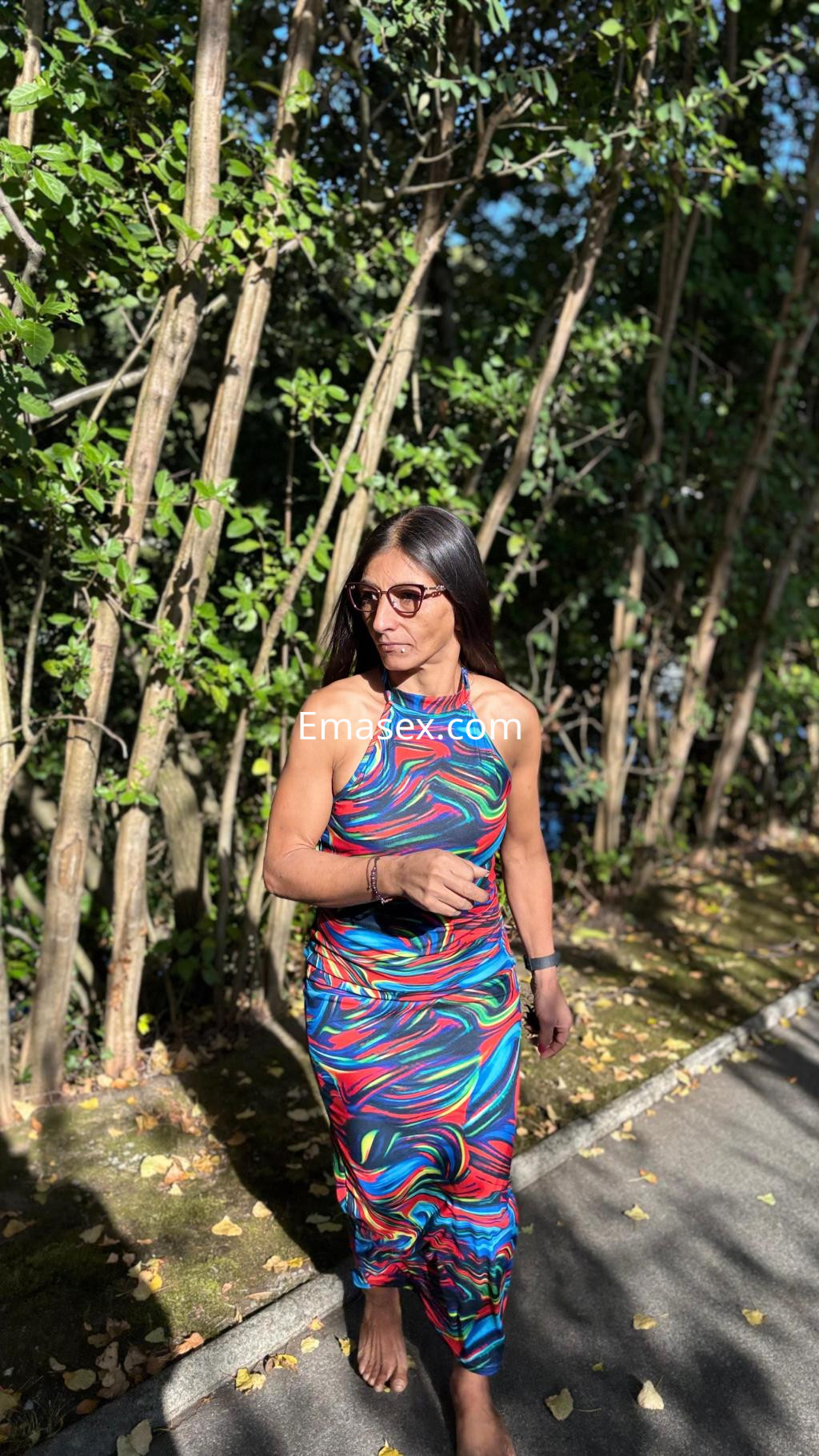 La puta camina por un sendero rodeado de árboles, luciendo un vestido colorido y gafas de sol en Irún.