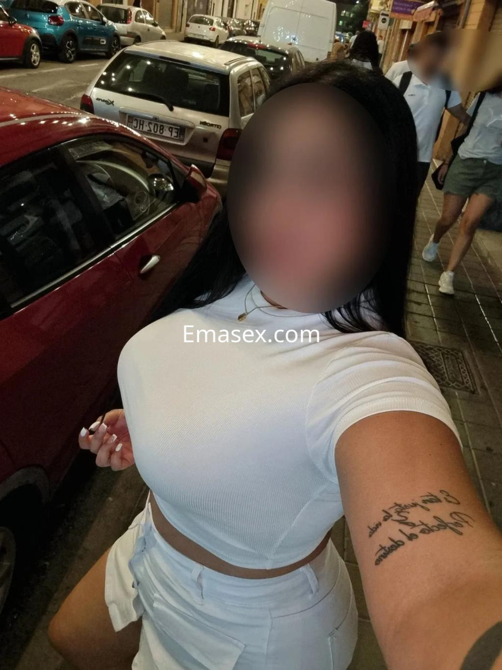 Puta en Alicante viste una blusa blanca ajustada y shorts, posando en una calle con coches de fondo.