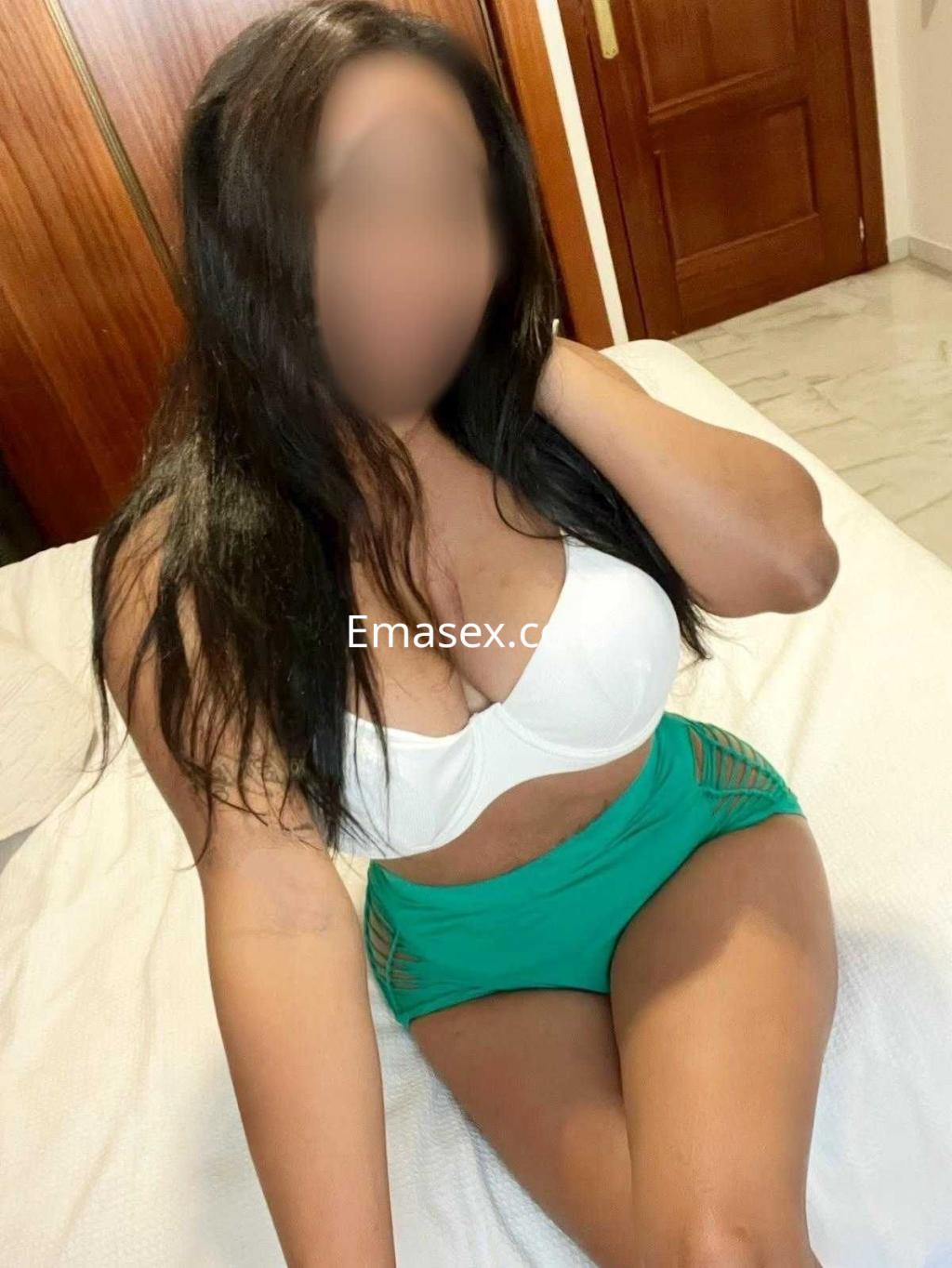 La puta luce un sujetador blanco y una falda corta verde, sentada en una cama en Sevilla.