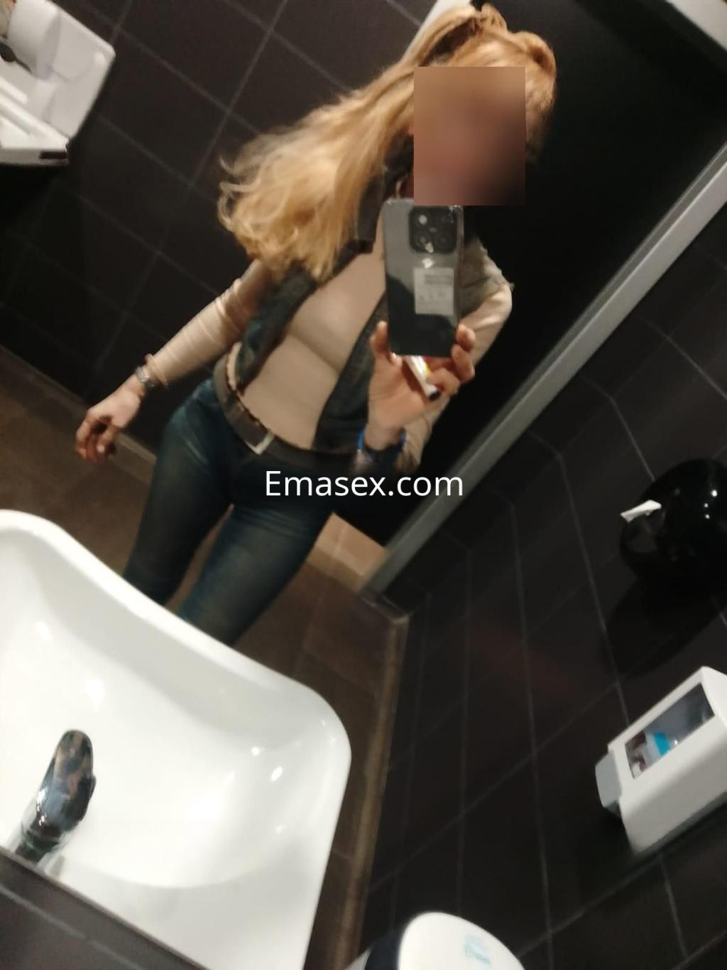Puta con cabello rubio y largo, viste una blusa clara y jeans ajustados en un baño de Málaga.