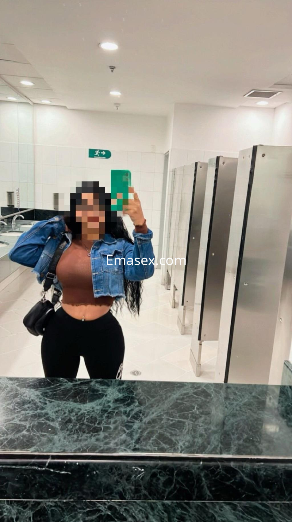 Puta en Málaga, con cabello rizado y largo, viste un top marrón y leggings negros, posando en un baño.