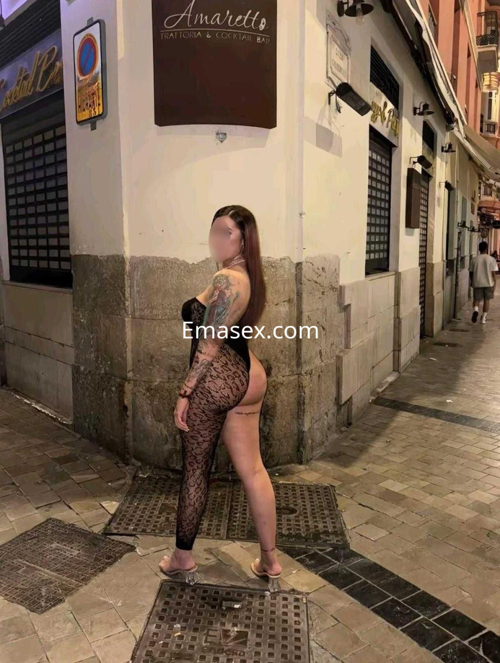 Puta en Granada, luciendo un body negro de encaje que resalta su figura y tatuajes en una calle iluminada.