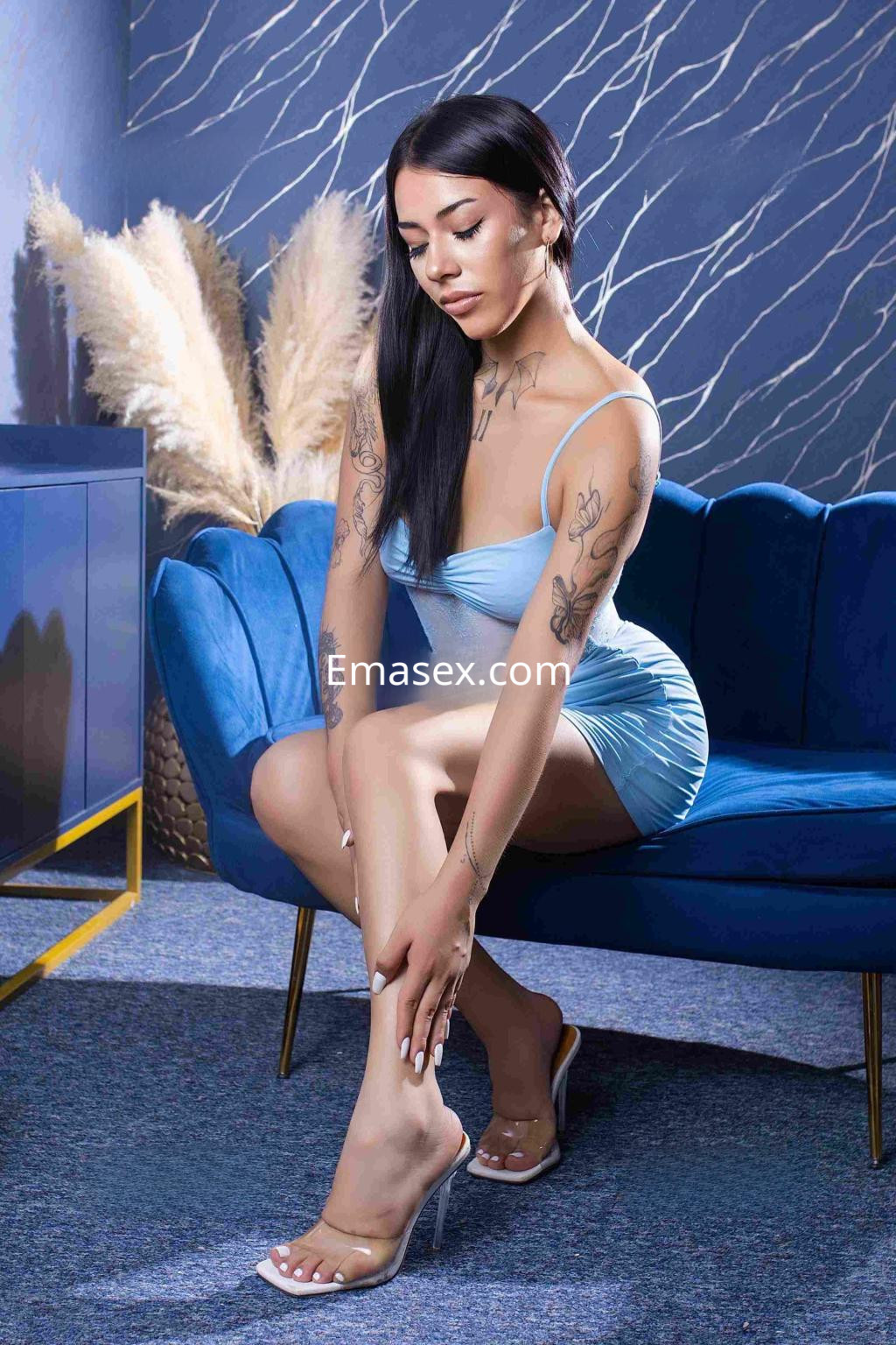 La puta está sentada en un sofá azul, vistiendo un vestido ajustado de color celeste y luciendo sus tatuajes en Barcelona.
