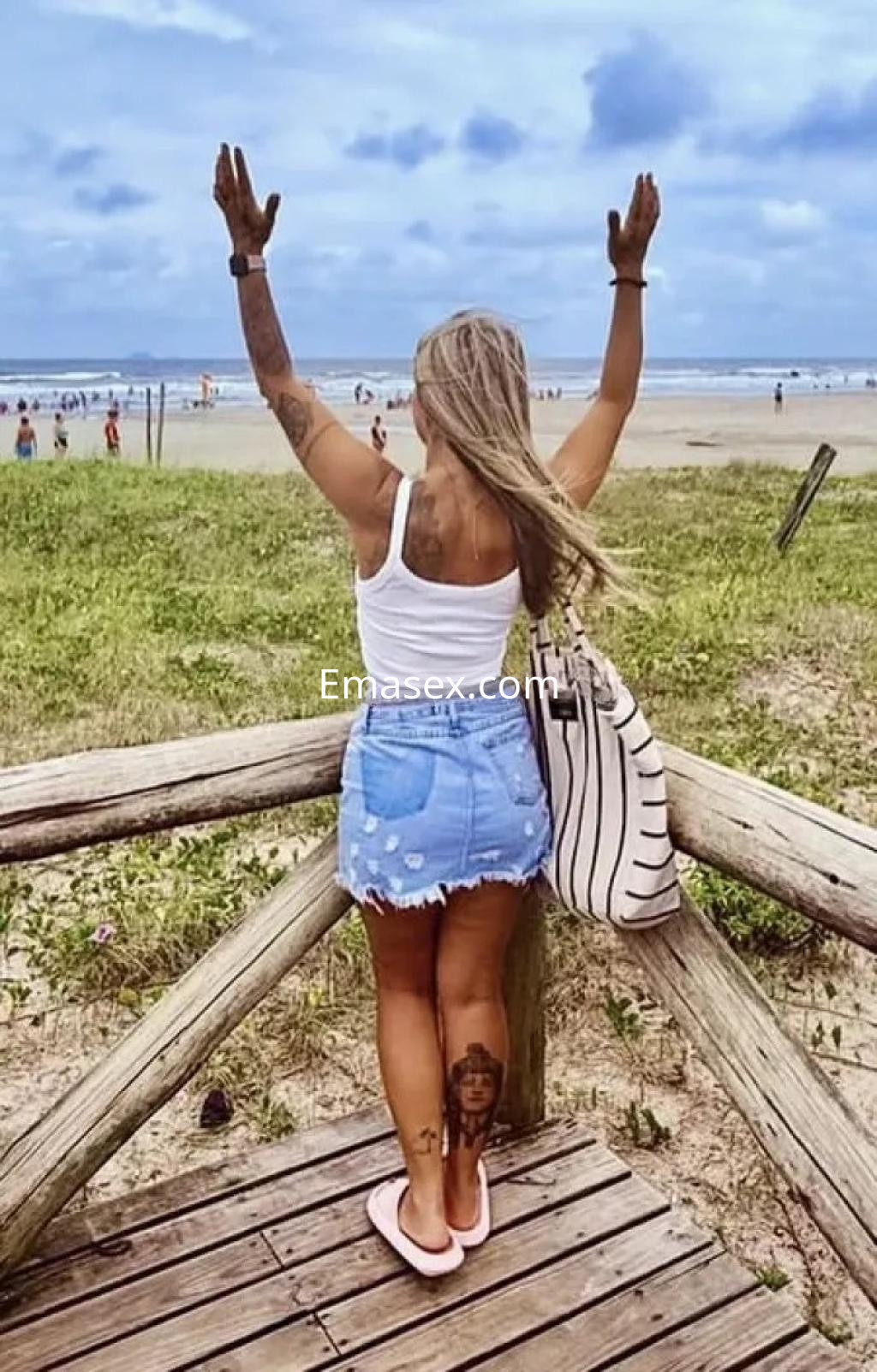 Escort de Coruña con cabello rubio, vistiendo camiseta blanca y falda de mezclilla, de pie en un entorno playero.