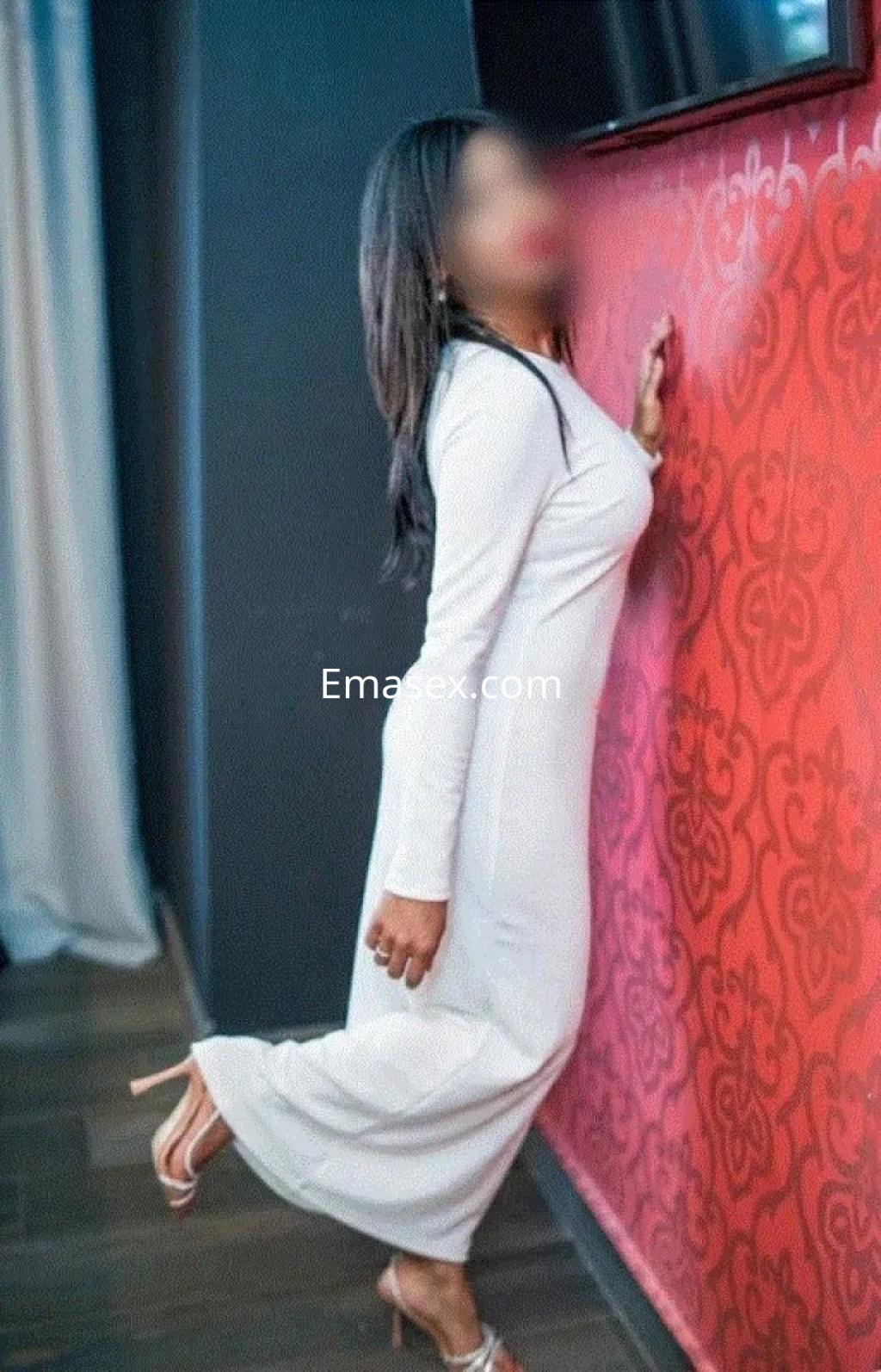 Escort en Alicante, luciendo un vestido largo blanco y posando con una pierna levantada junto a una pared decorativa.