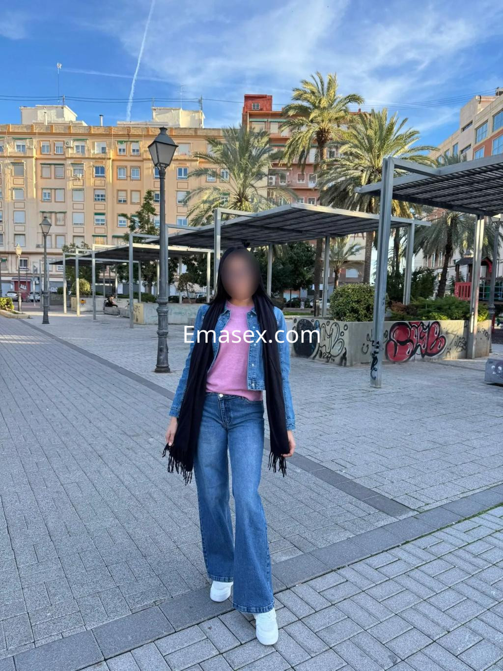 Escort en Valencia viste un suéter rosa y jeans anchos, posando en una plaza con palmeras y edificios.