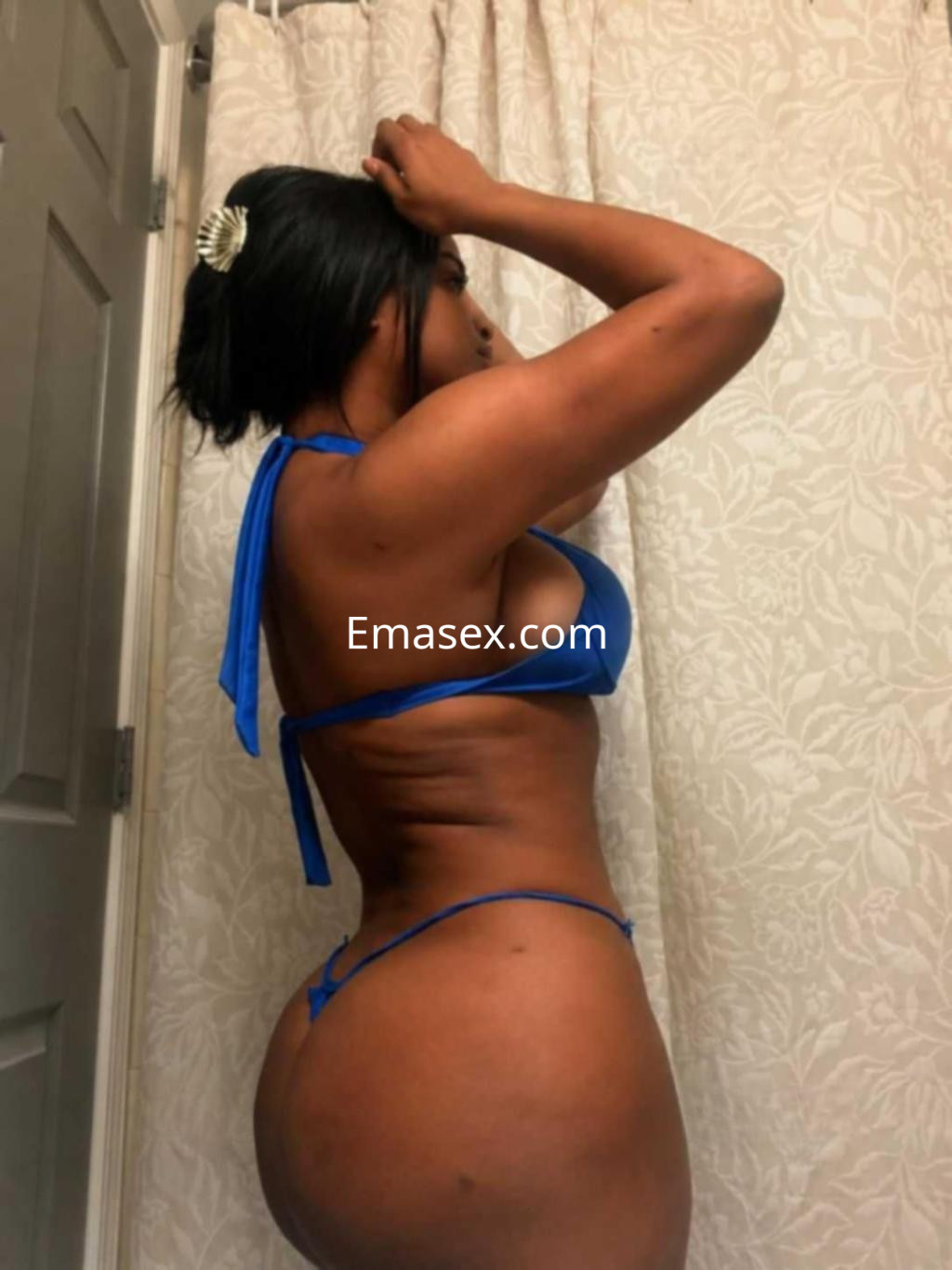 Escort en Barcelona con cabello oscuro, usando un bikini azul, posando de perfil frente a un fondo neutro.