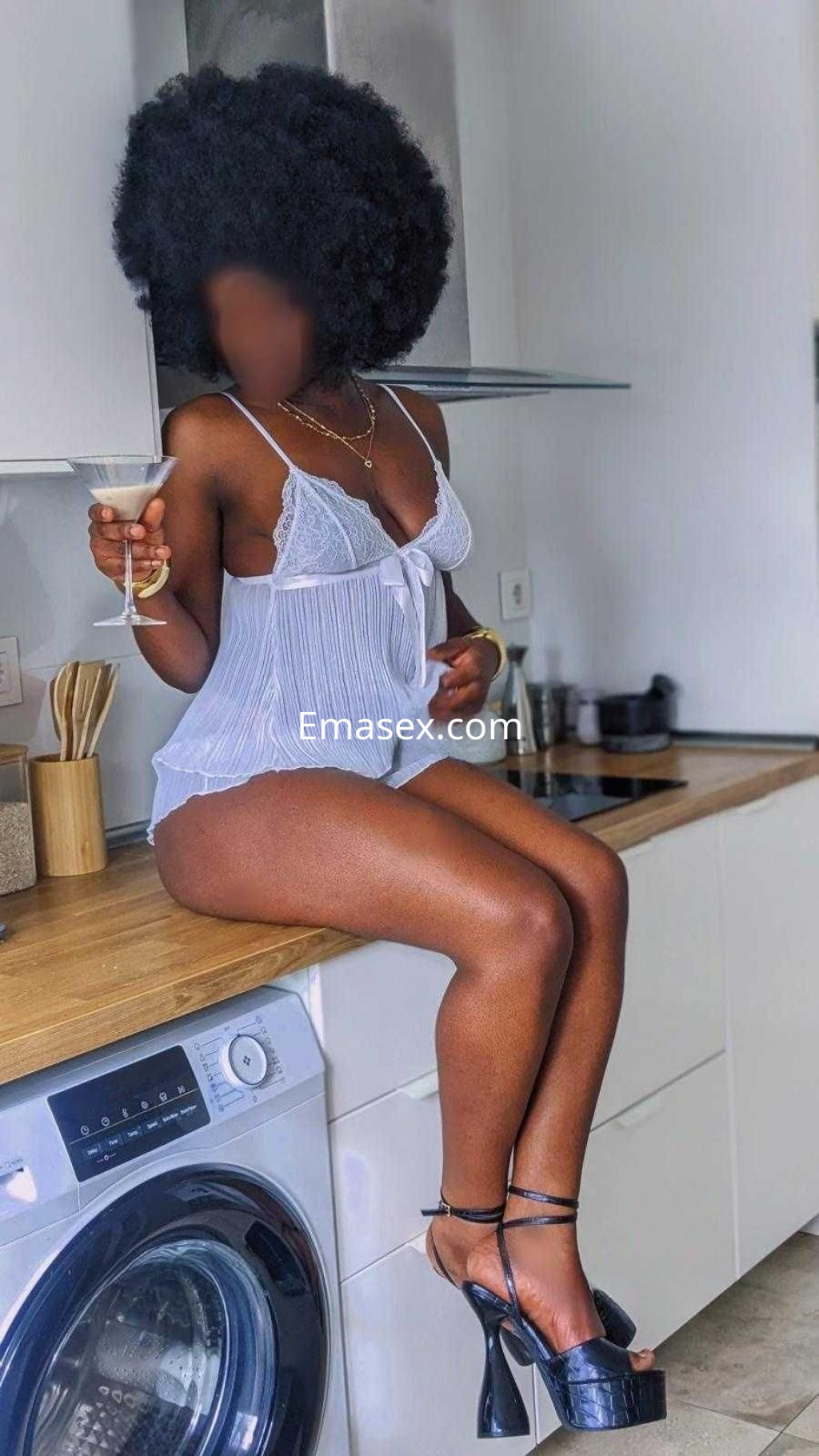 Puta sentada en la encimera de una cocina en Barcelona, luciendo un conjunto blanco y tacones altos mientras sostiene una copa.