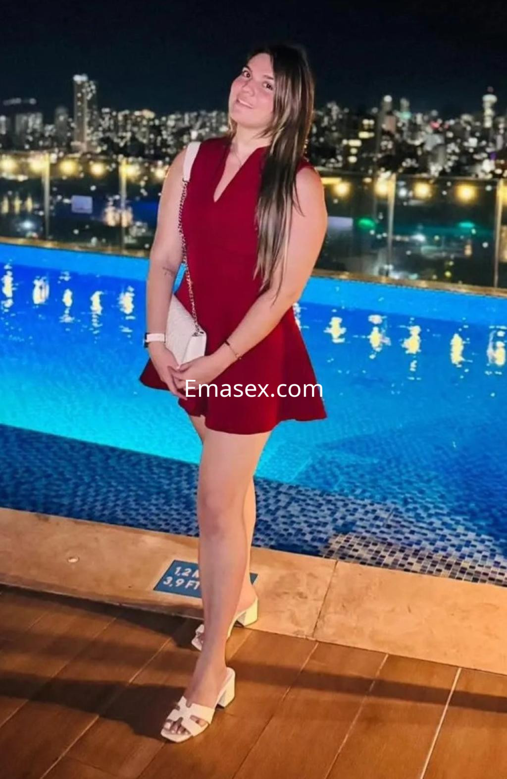 Escort en Madrid, luciendo un vestido rojo, de pie junto a una piscina iluminada y con la ciudad de fondo.
