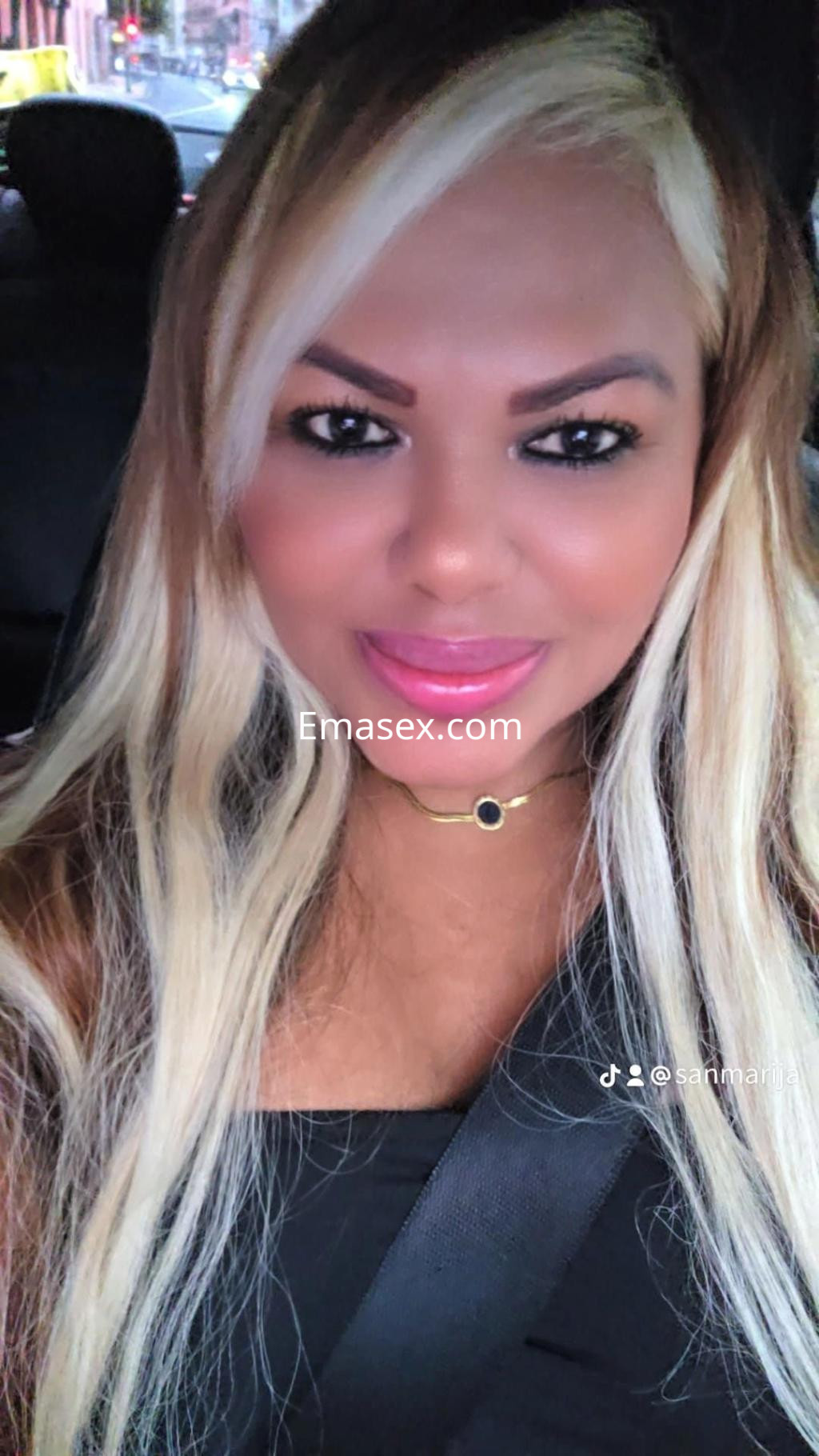 Escort en Sevilla con cabello largo y rubio, luciendo un top negro, sentada en un coche y sonriendo a la cámara.