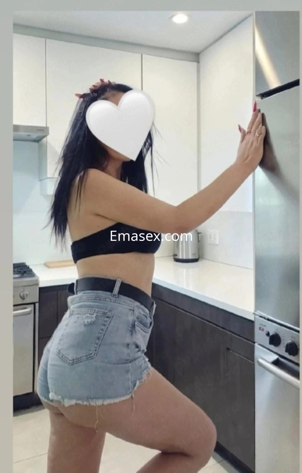 Escort en Gijón posando en una cocina moderna, luciendo un top negro y shorts de mezclilla.