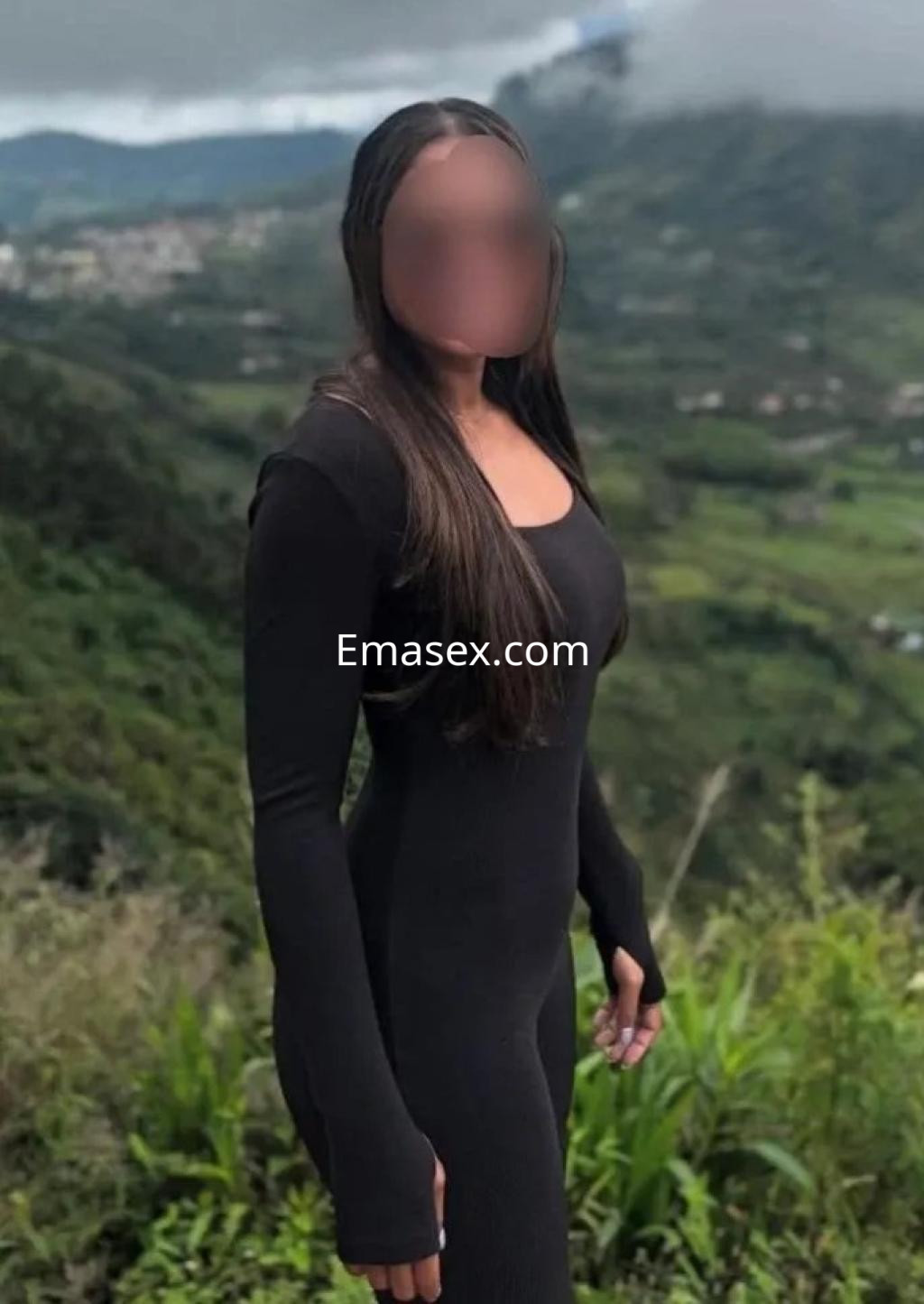 Escort en Oviedo, de pie en un paisaje verde, viste un mono negro que resalta su figura y tiene el cabello largo.