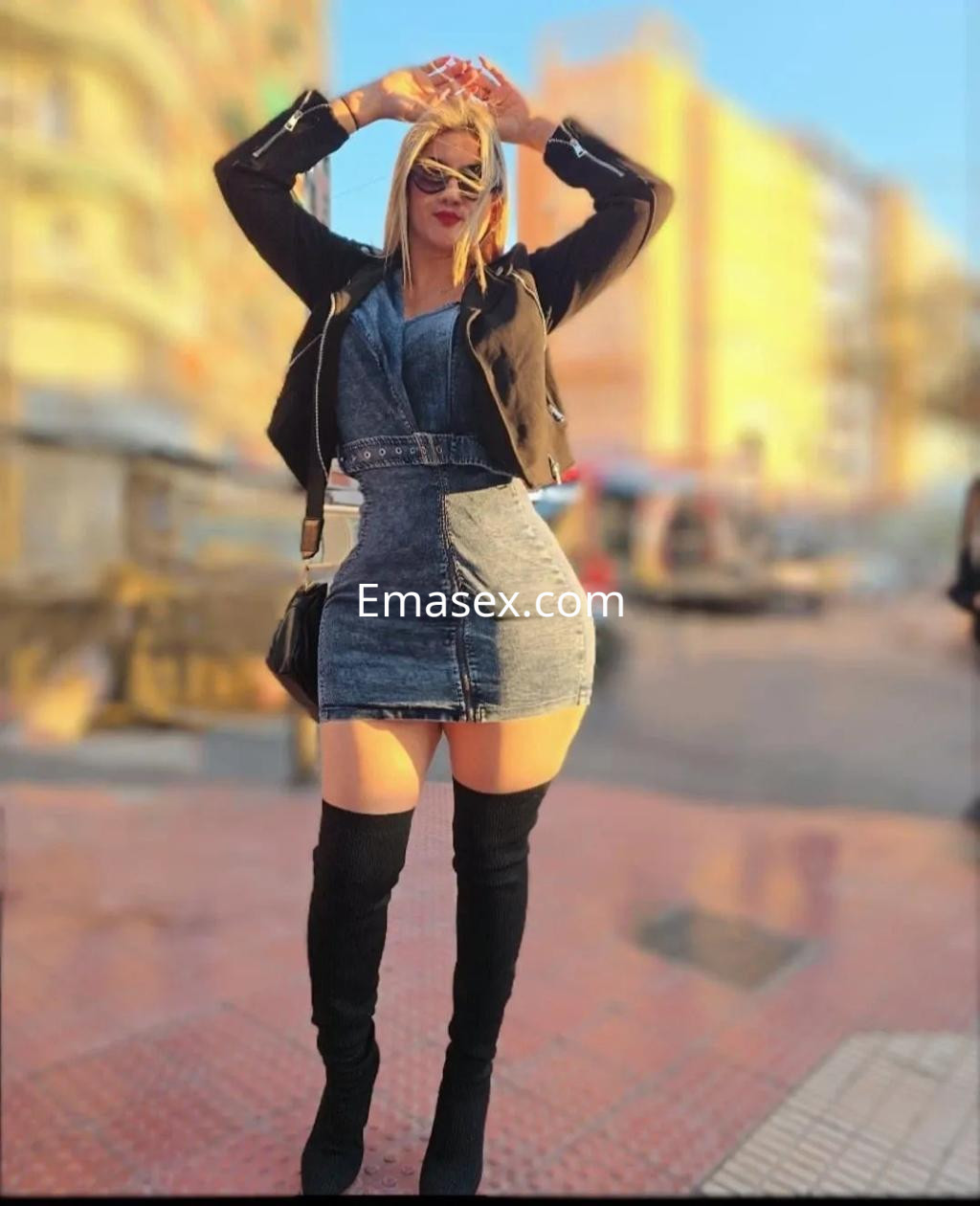 Puta en Alicante viste un ajustado vestido de mezclilla, con botas altas y gafas de sol, posando al aire libre.
