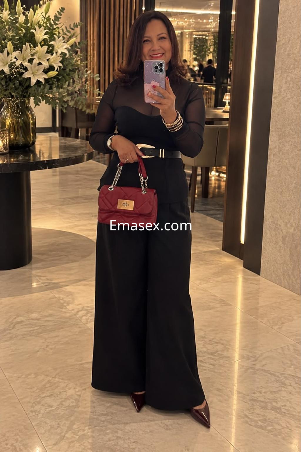 Escort de cabello castaño oscuro y blusa transparente, posando con pantalones negros y bolso rojo en un elegante entorno.