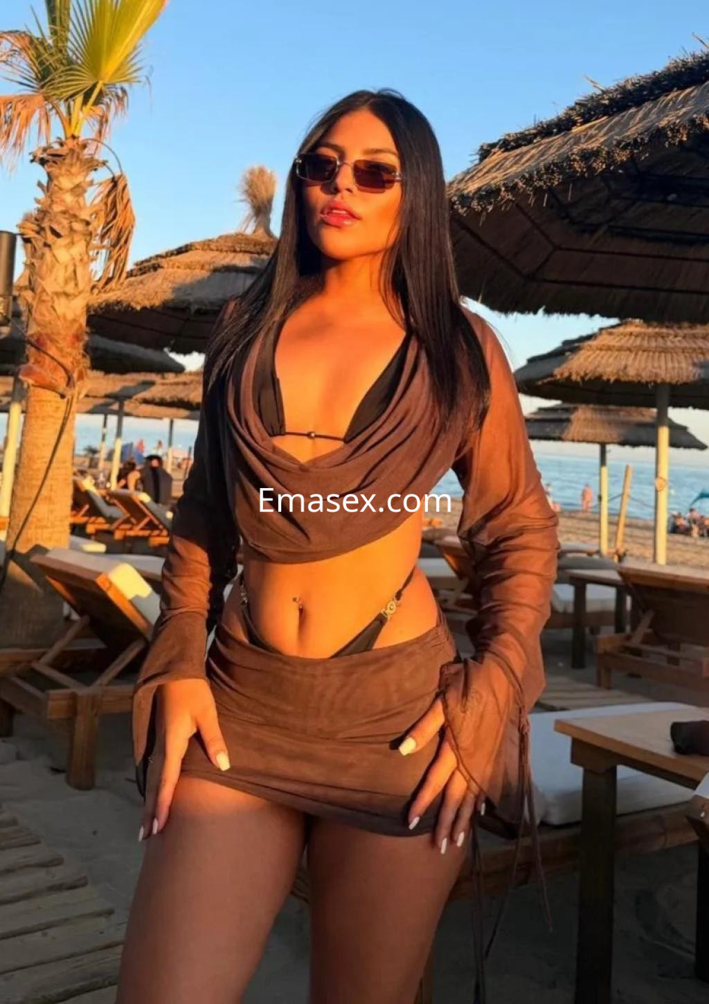La puta en Zaragoza lleva un conjunto marrón claro con un bikini negro, posando bajo la luz del sol en la playa.