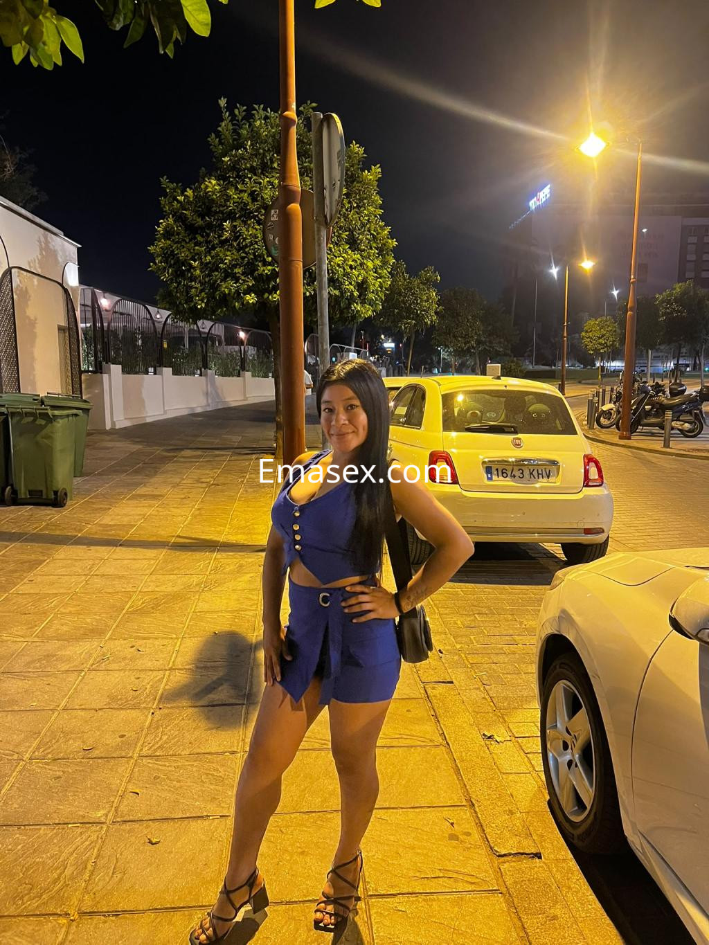 Escort en Sevilla, de pie en la calle por la noche, con un vestido azul y tacones, sonriendo a la cámara.