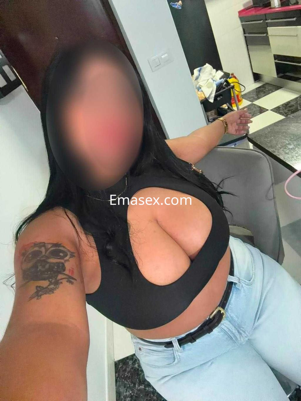 Escort en Cádiz con blusa negra de escote pronunciado y jeans claros, posando en una cocina moderna.