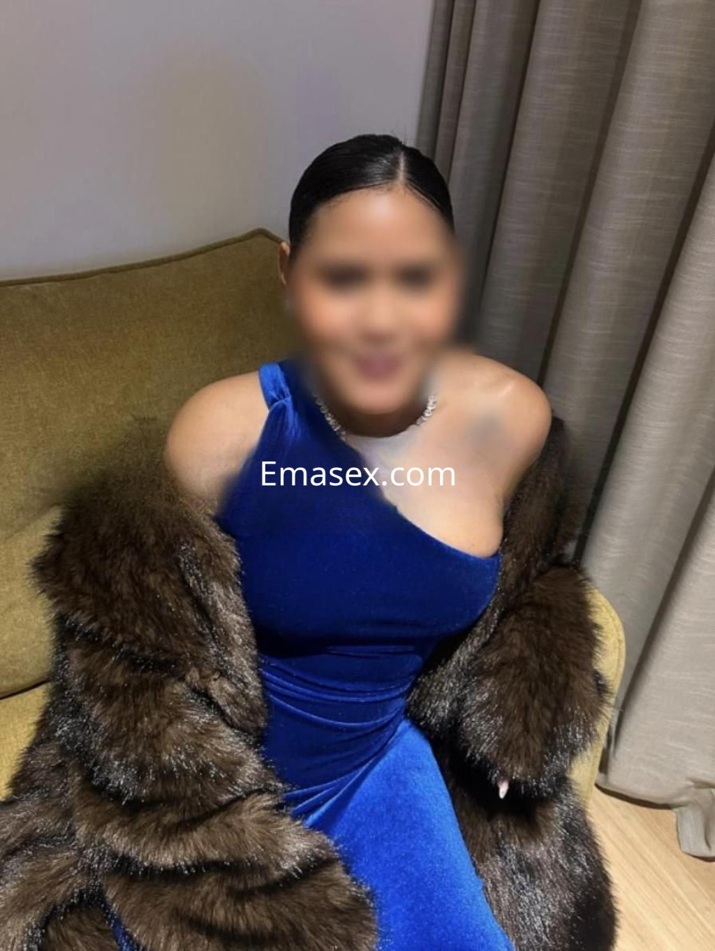 Puta con vestido azul de una sola manga y abrigo de piel, sentada en un sofá en Madrid.