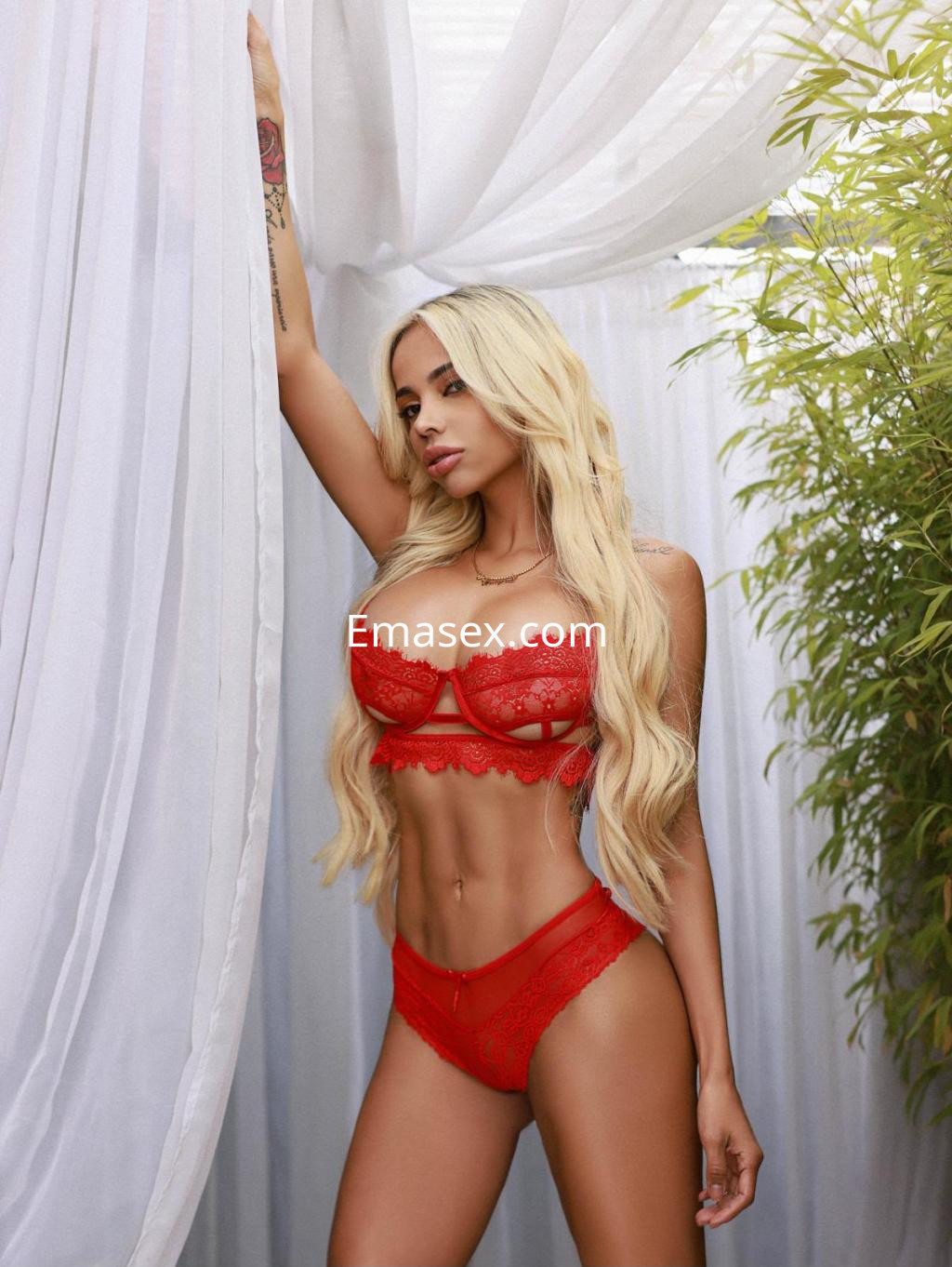 Escort en Ibiza, posando en lencería roja de encaje, con cabello rubio y fondo de cortinas blancas.