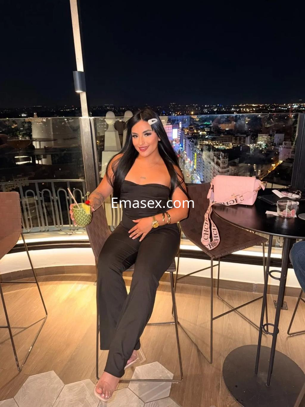 La escort en Valencia luce un conjunto negro ajustado, sentada en una terraza con vista nocturna, sosteniendo una bebida.