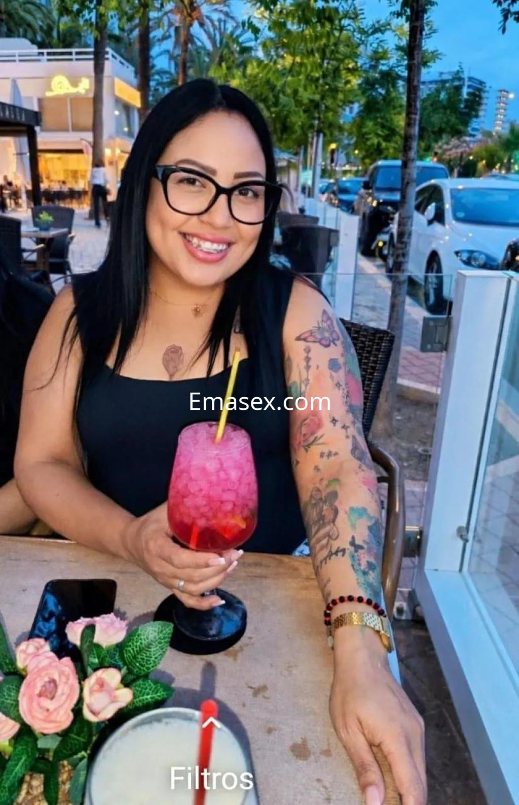 La escort sonríe mientras sostiene un cóctel colorido en una terraza de Palma de Mallorca, luciendo un top negro y tatuajes.