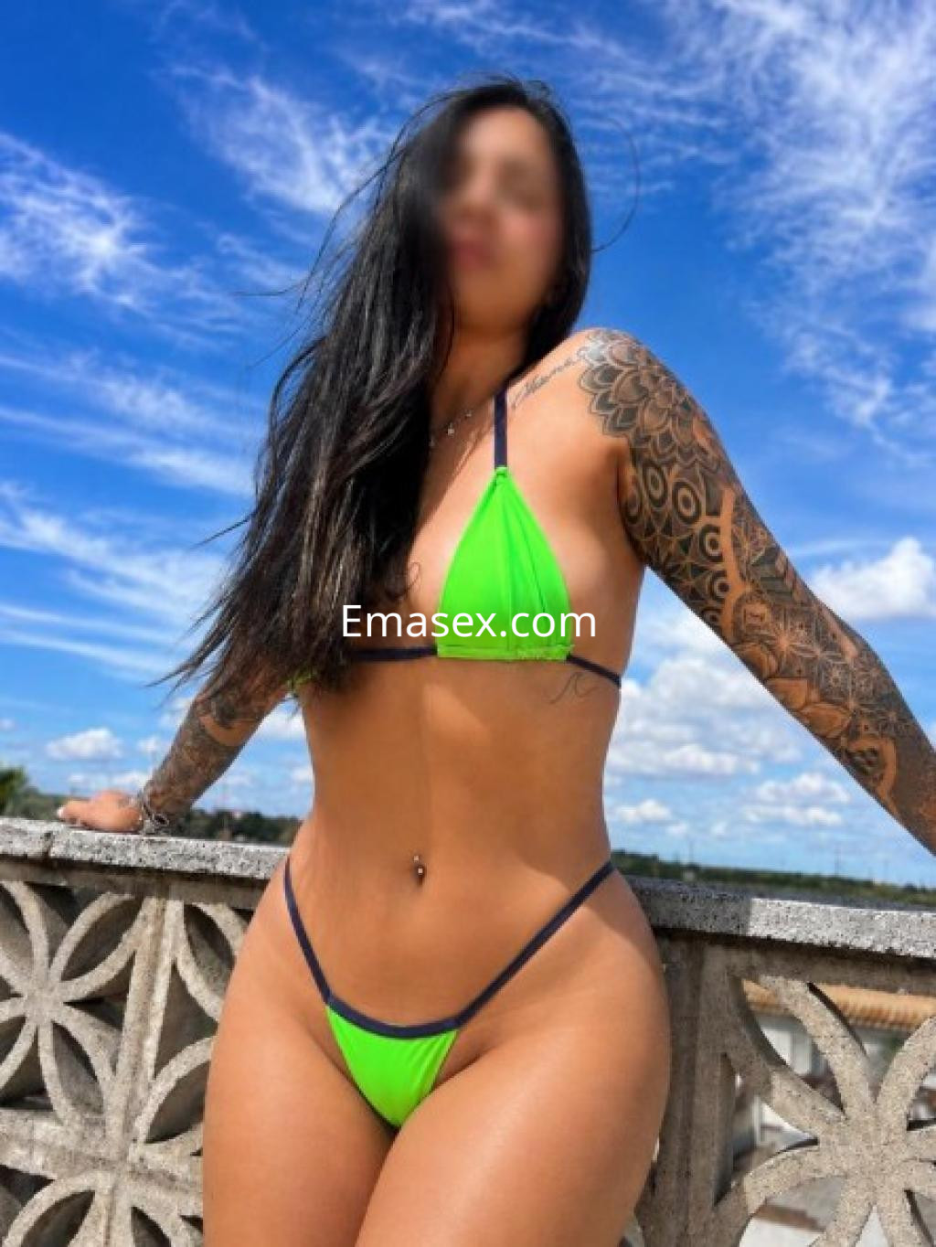 Escort en Cádiz con cabello largo y oscuro, luciendo un bikini verde y negro, posando con fondo de cielo despejado.