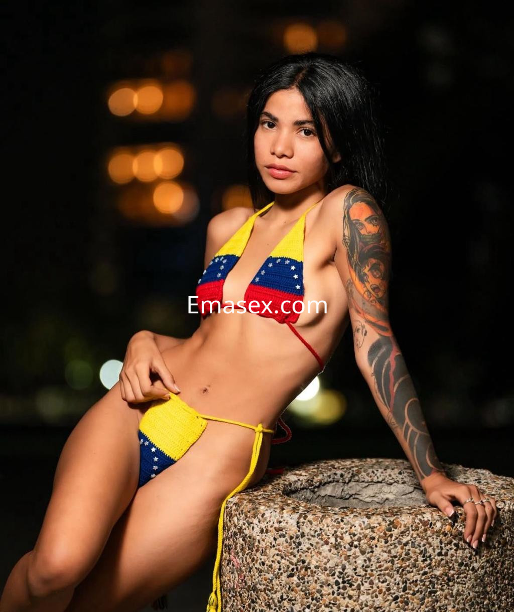 Escort en Donosti luciendo un bikini colorido y un tatuaje en el brazo, posando de manera relajada sobre una piedra.