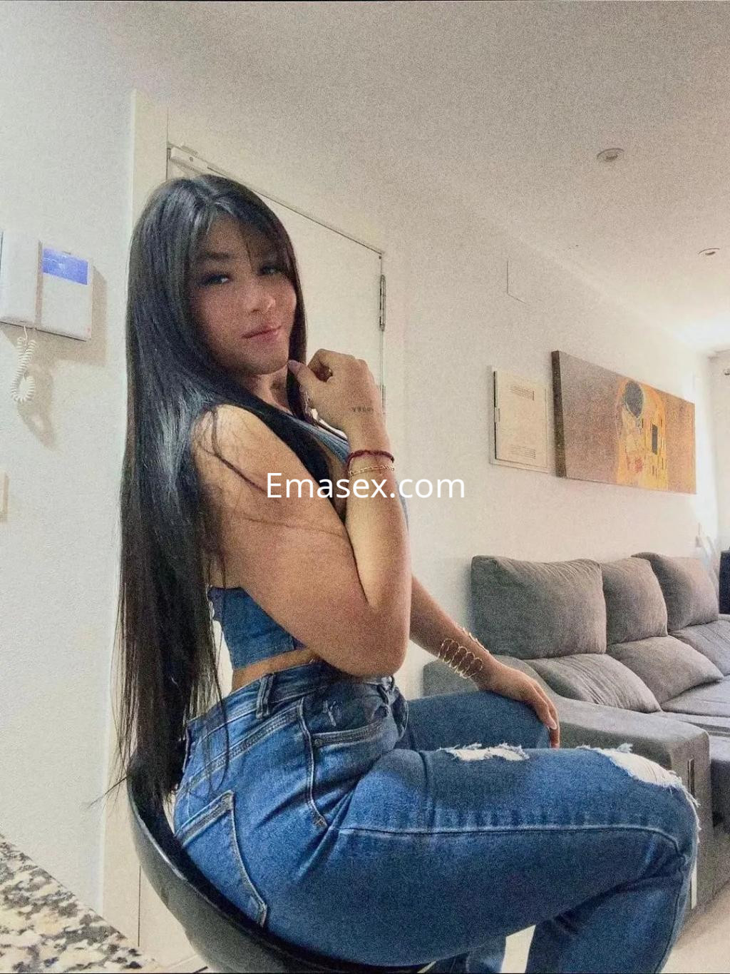 Escort en Coruña con cabello largo y lacio, vistiendo un top azul y jeans ajustados, posando sobre una silla.