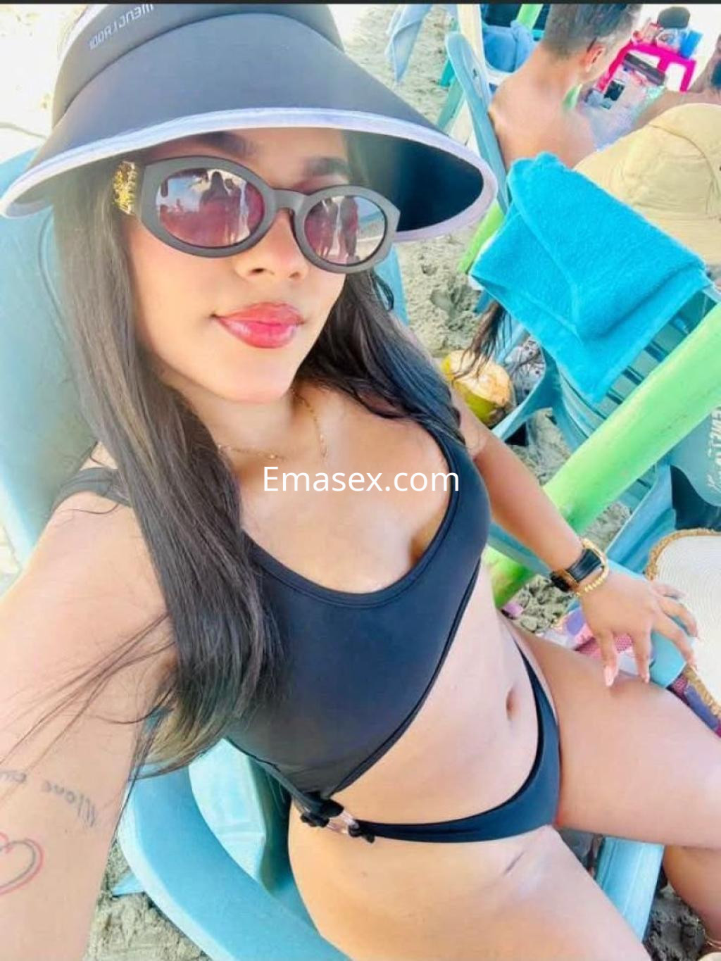 Puta en Valencia con bikini negro, gafas de sol y sombrero, posando en una playa con sillas de verano.