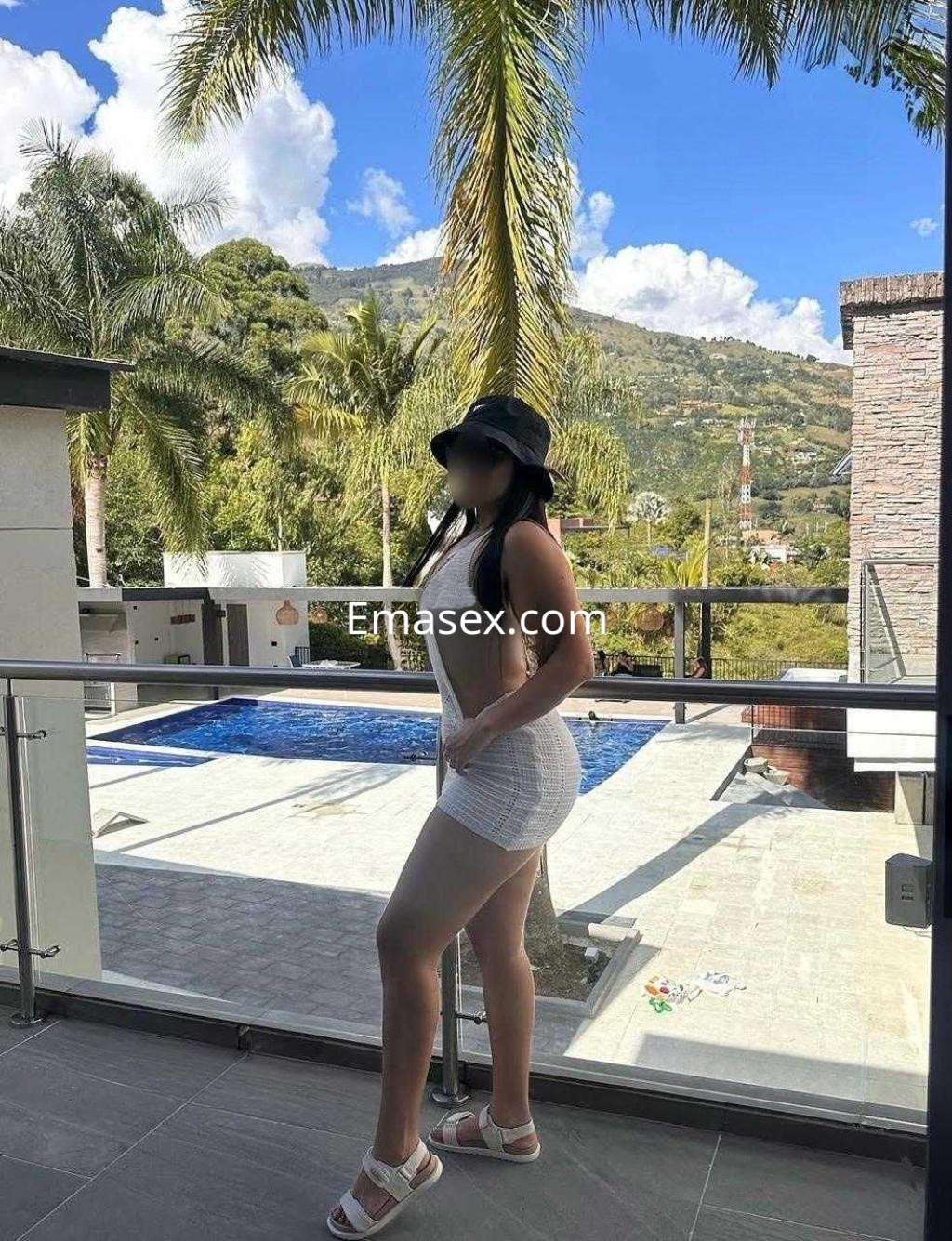Puta de pie frente a una piscina en Valencia, luciendo un vestido blanco y un sombrero negro, con un paisaje verde de fondo.