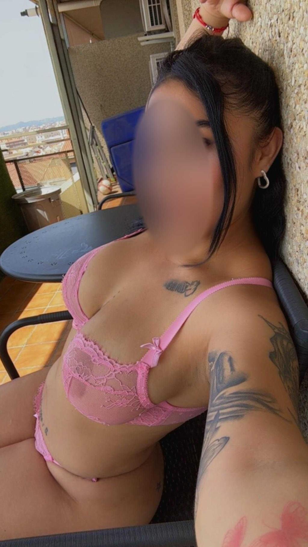 Puta en Valencia posando con lencería rosa en un balcón, mostrando su tatuaje y una mirada seductora.