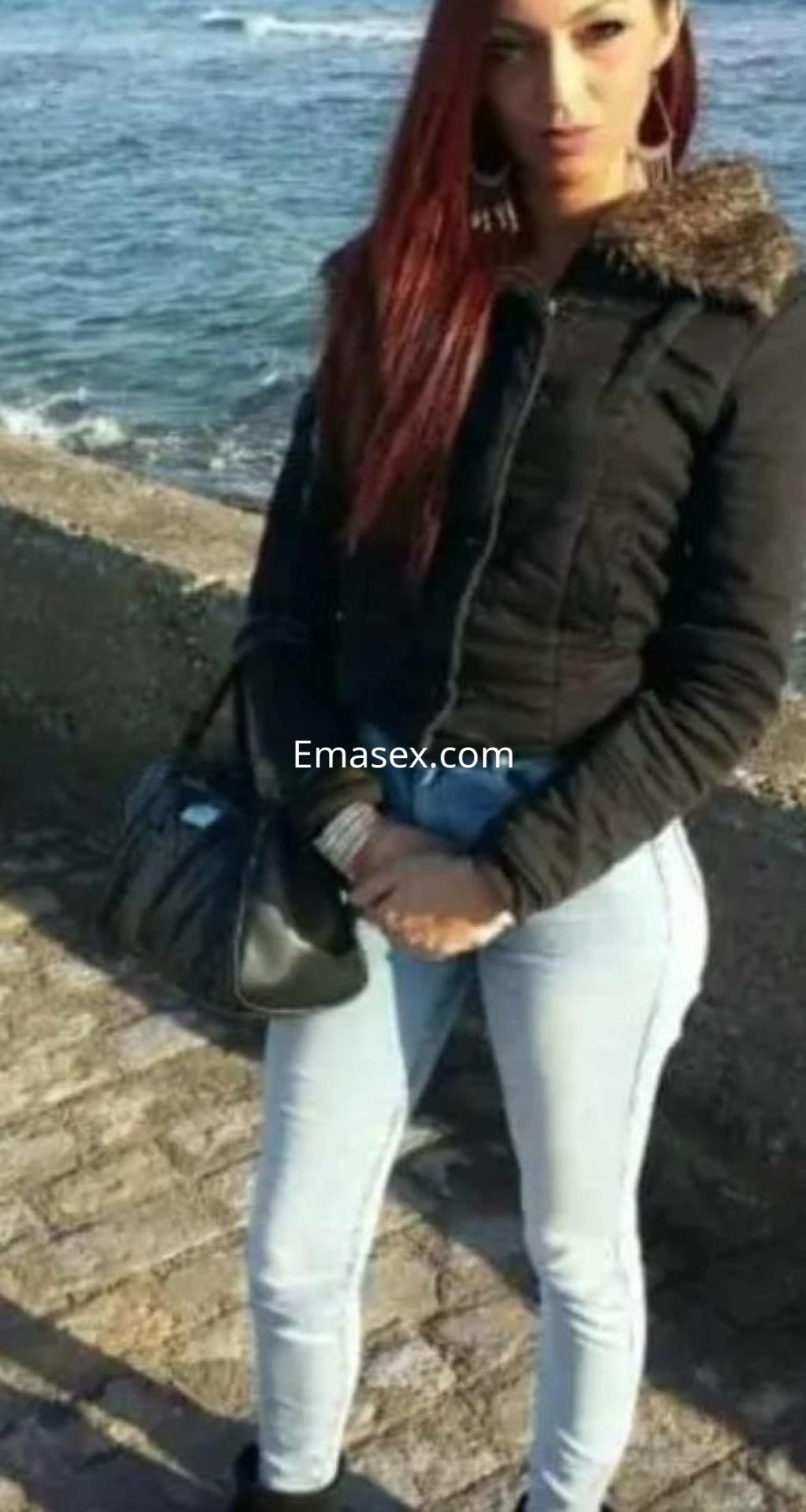 Puta en Oviedo con cabello largo y rojo, vestida con chaqueta negra y jeans ajustados, de pie junto al mar.