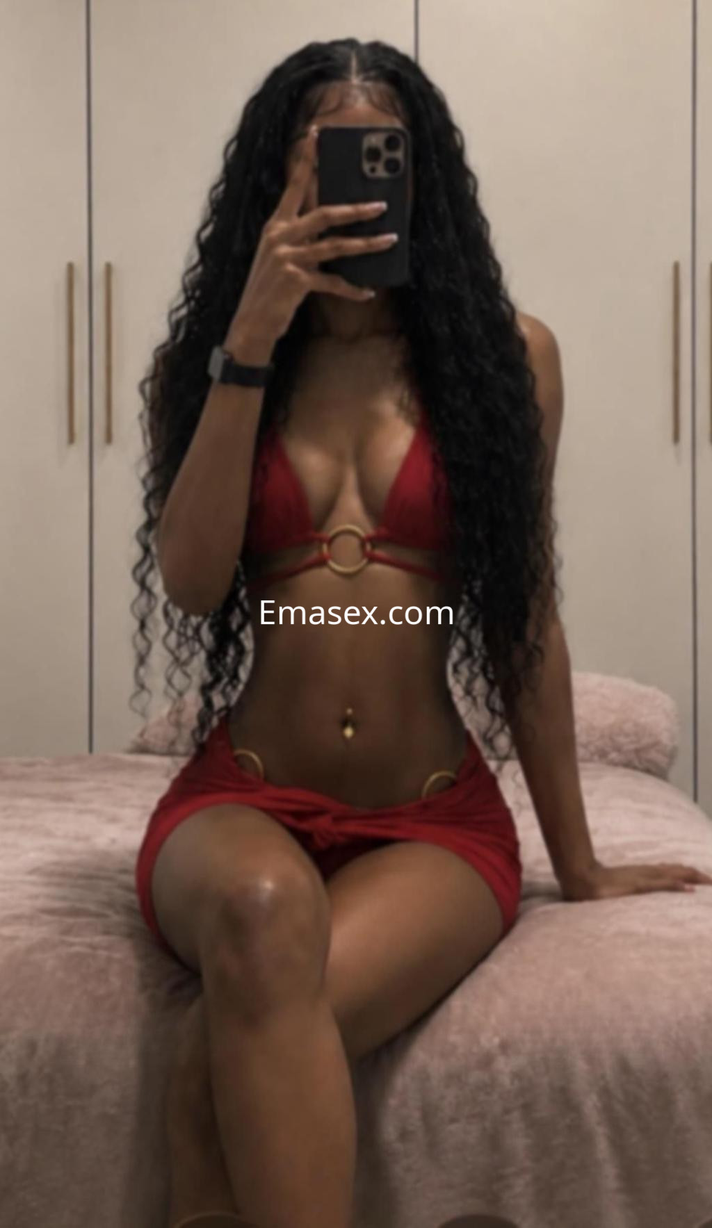 Puta en Barcelona con cabello rizado y largo, vistiendo un bikini rojo y una falda corta, posando en un entorno interior.