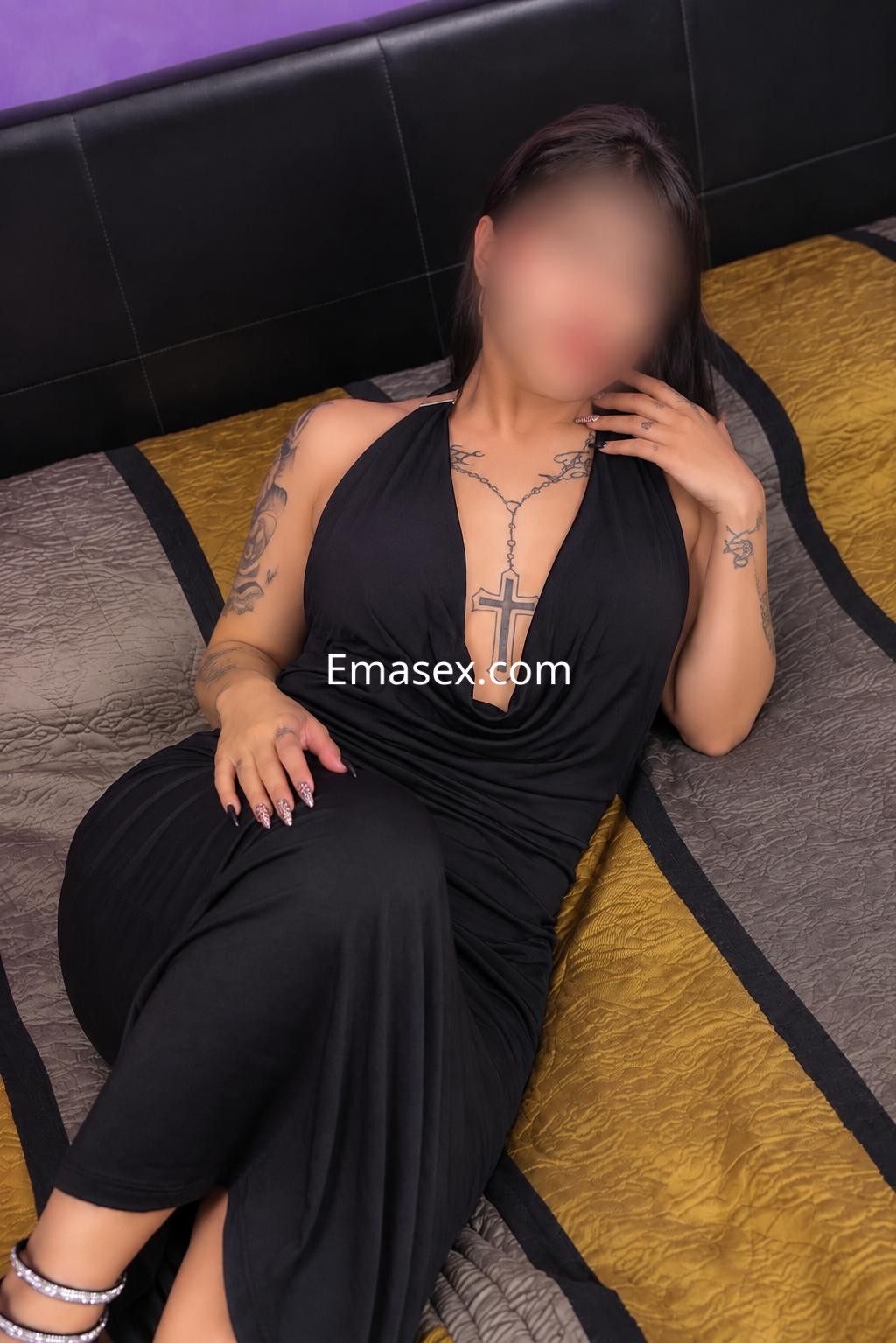 Puta en Vitoria lleva un vestido negro con escote pronunciado y tatuajes visibles, posando sobre la cama.