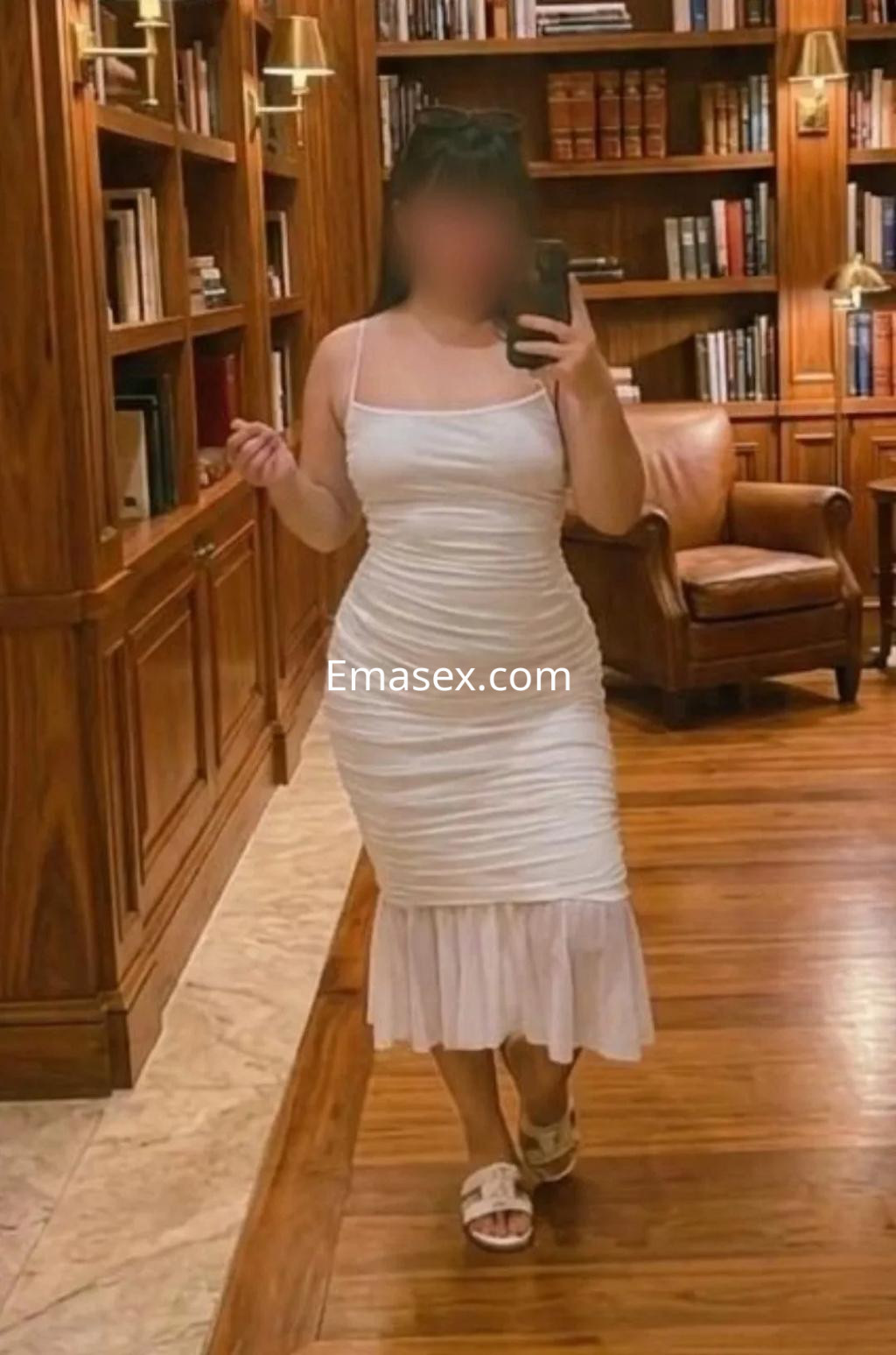 Escort en Alicante, usando un vestido blanco ajustado y sandalias, en un elegante entorno de biblioteca.