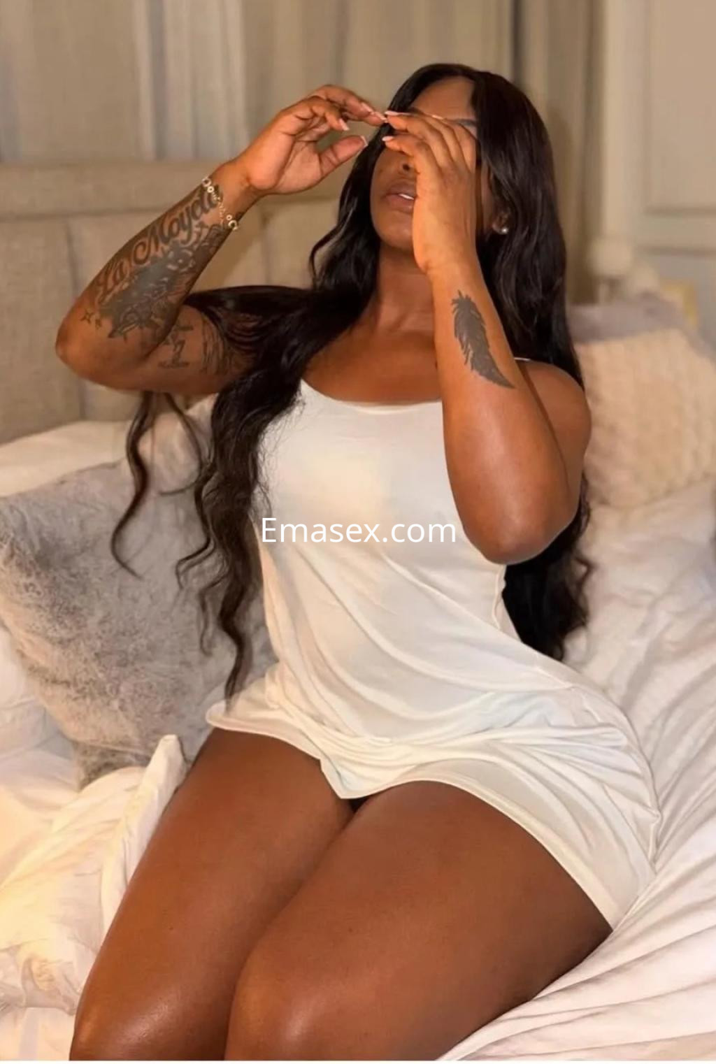 La escort en Zaragoza posa sentada en la cama, vistiendo una camiseta blanca sin mangas y mostrando su cabello largo y ondulado.
