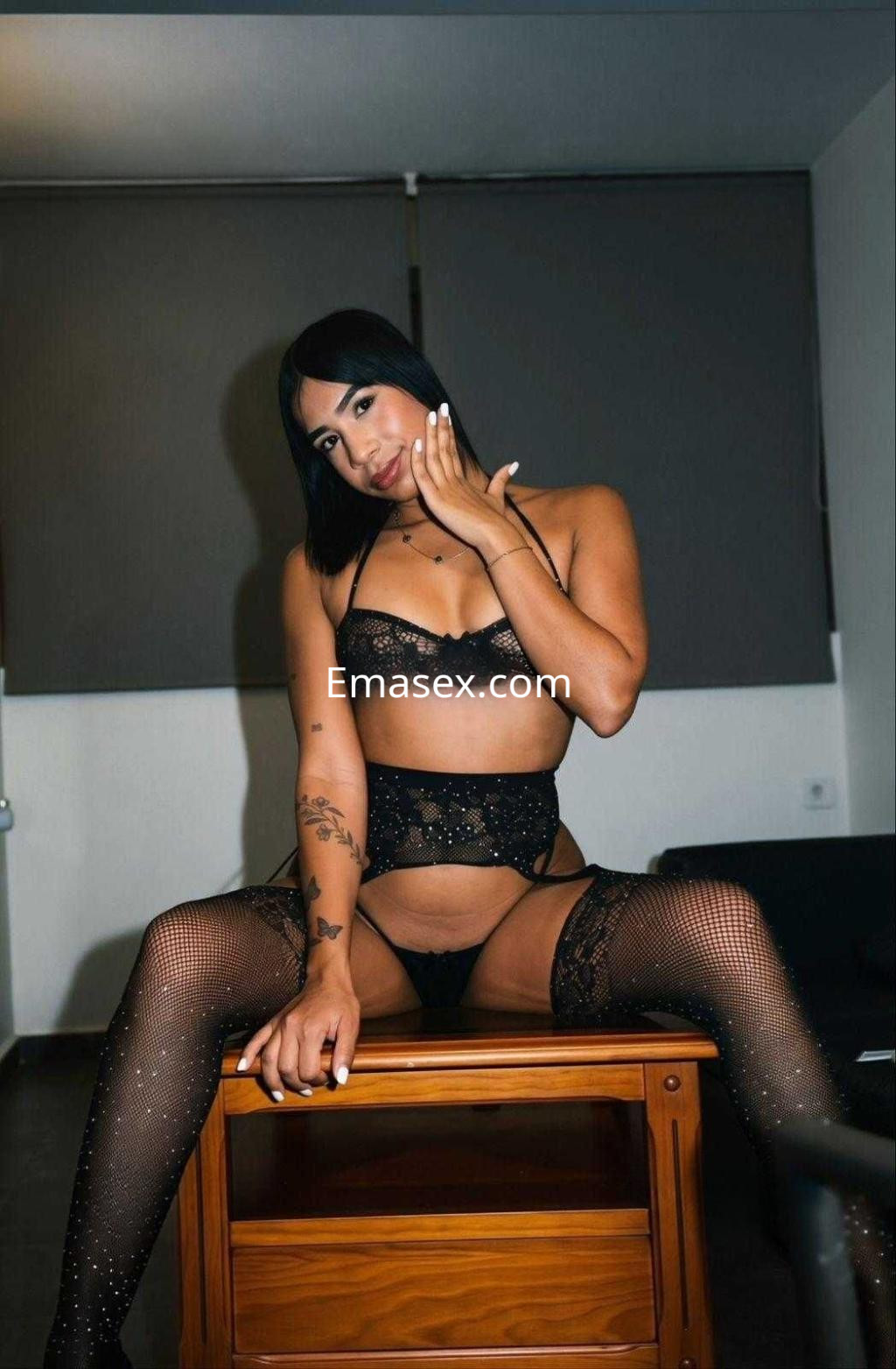 Escort en Alicante posando sentada en una mesa, con lencería negra y medias de red, cabello lacio y corto.
