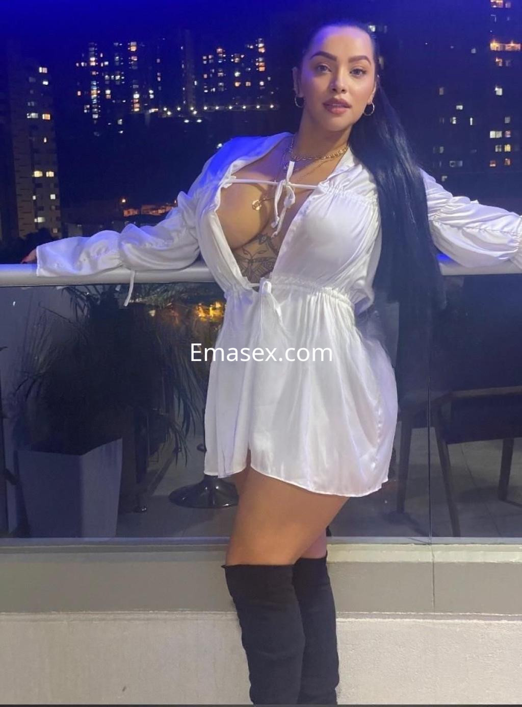 Escort en Girona con cabello largo y oscuro, vistiendo una camisa blanca abierta, posando en un balcón nocturno.