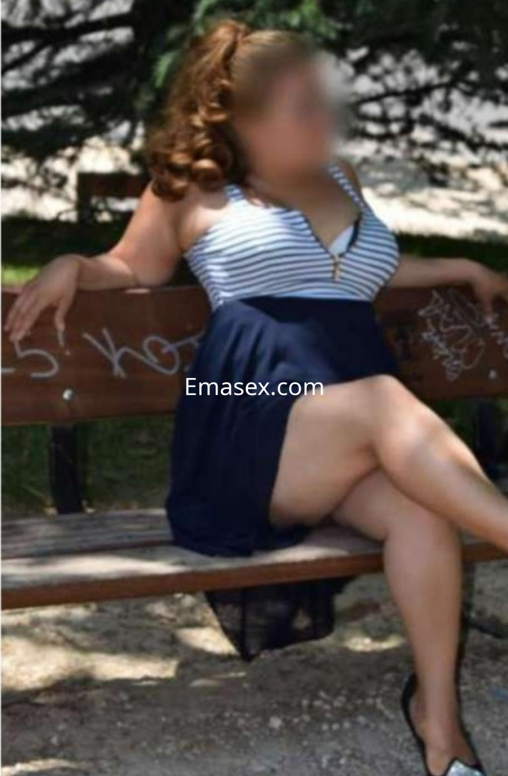 Escort sentada en un banco de parque, vistiendo un vestido azul con rayas y sandalias, en Coruña.