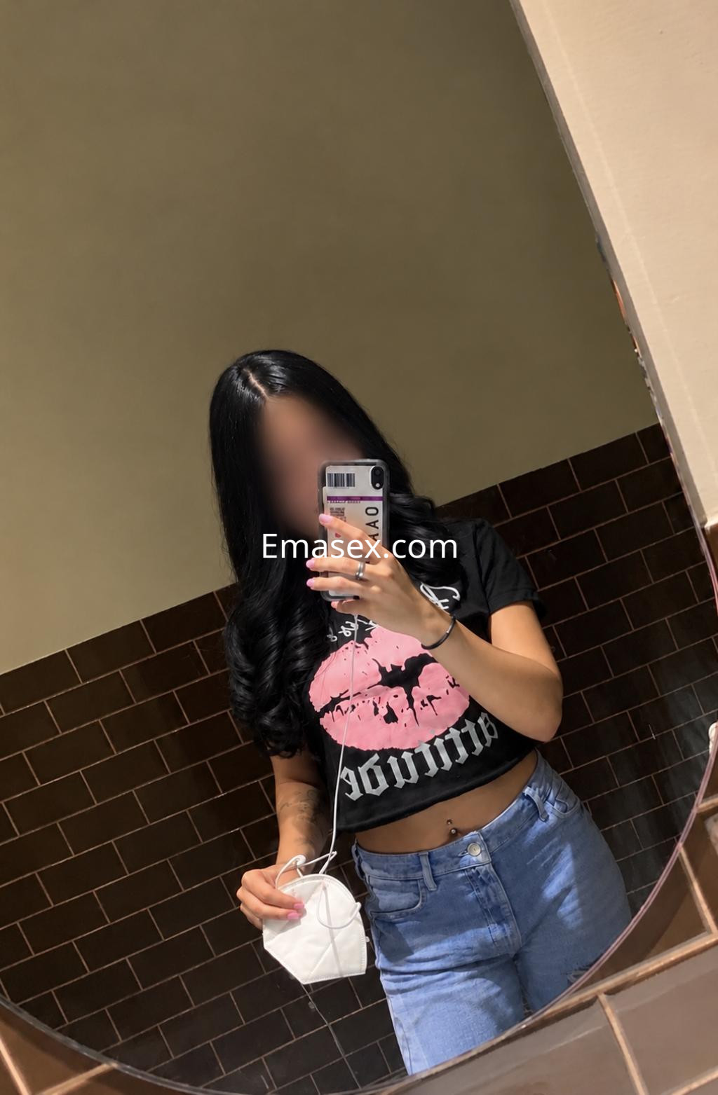 Puta en Santander, posando en un espejo con una camiseta negra y jeans claros, sosteniendo una mascarilla.