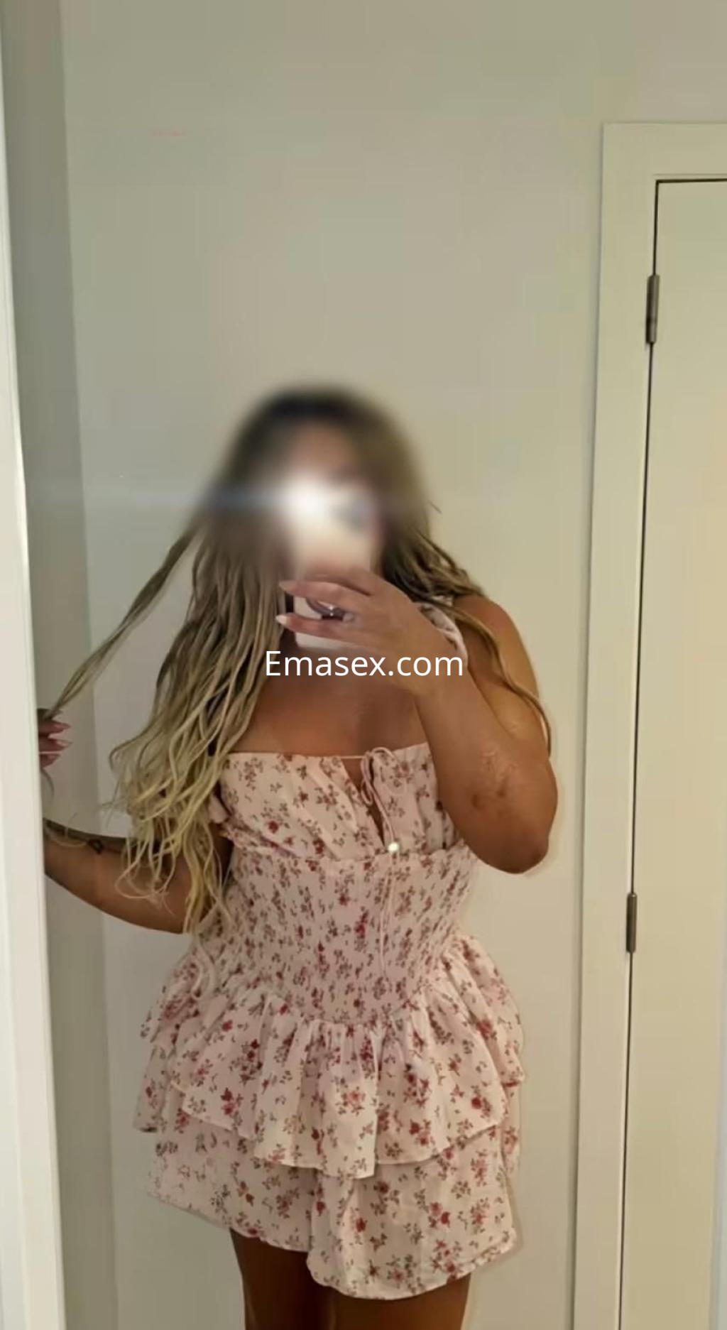 Puta en Valencia, luciendo un vestido corto de flores y mostrando su cabello largo y ondulado frente al espejo.