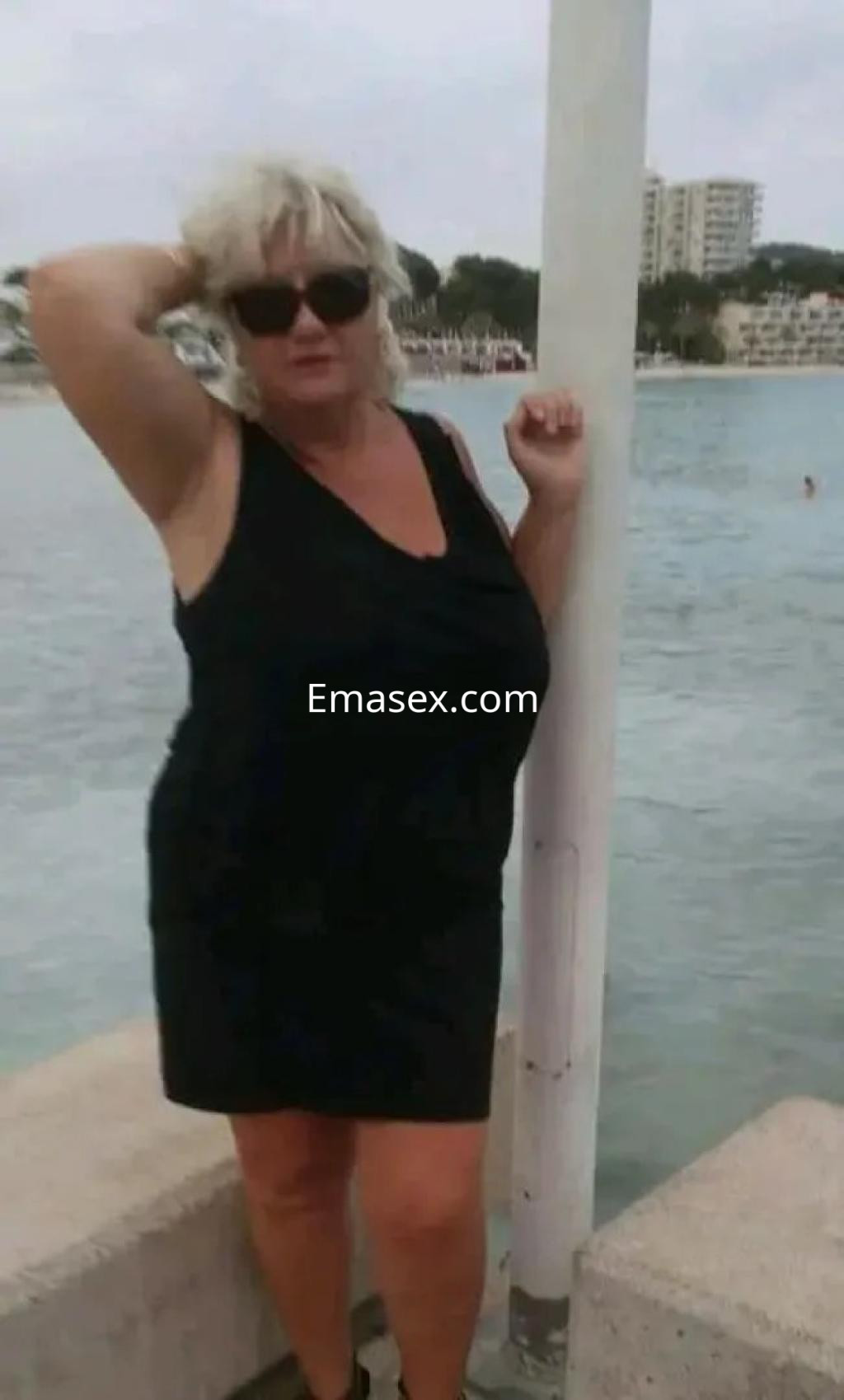 Puta con vestido negro y gafas de sol posando junto al agua en Palma de Mallorca, mostrando una actitud relajada.