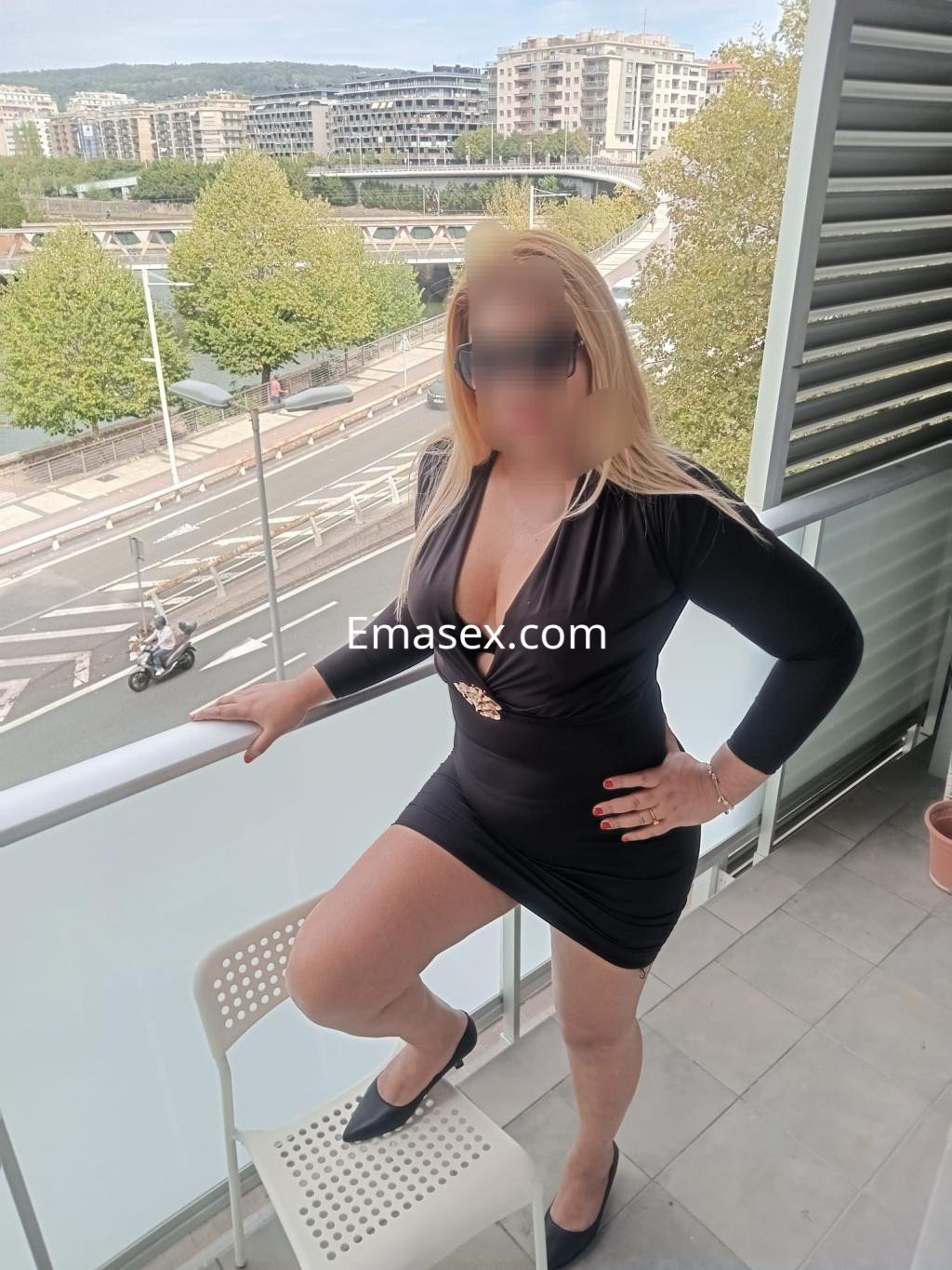 La escort posa con un vestido negro ajustado y tacones en un balcón en Donosti, con el cabello rubio suelto.