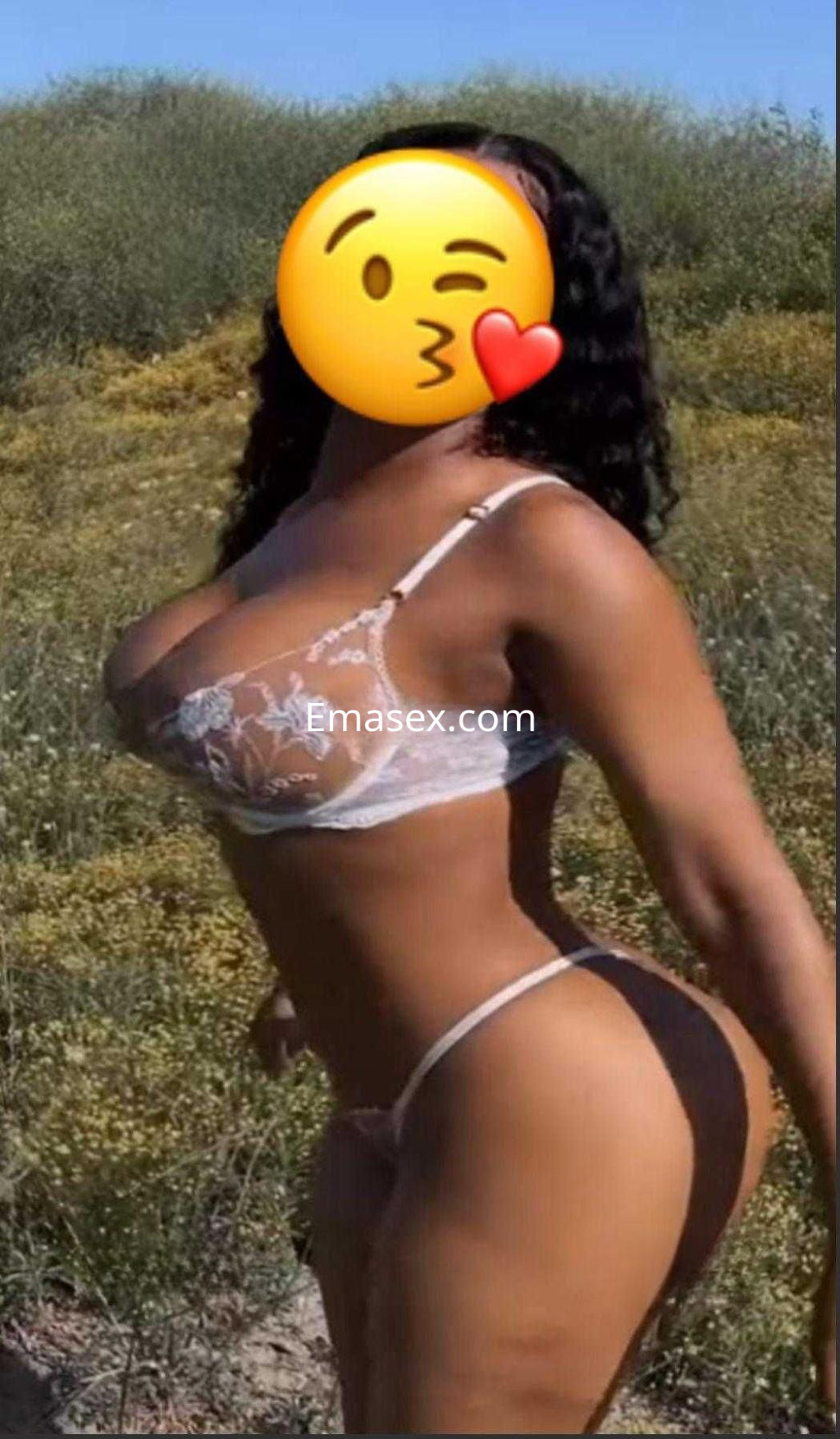 Escort en Málaga posando de perfil con lencería blanca, cabello rizado y un fondo natural.