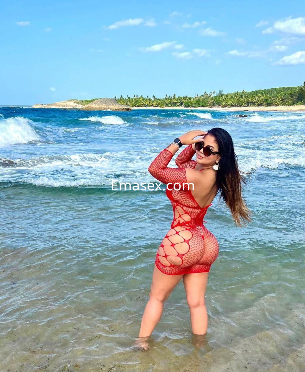 Puta con cabello largo y lacio, lleva un vestido de red rojo, posando en la playa con olas en Málaga.