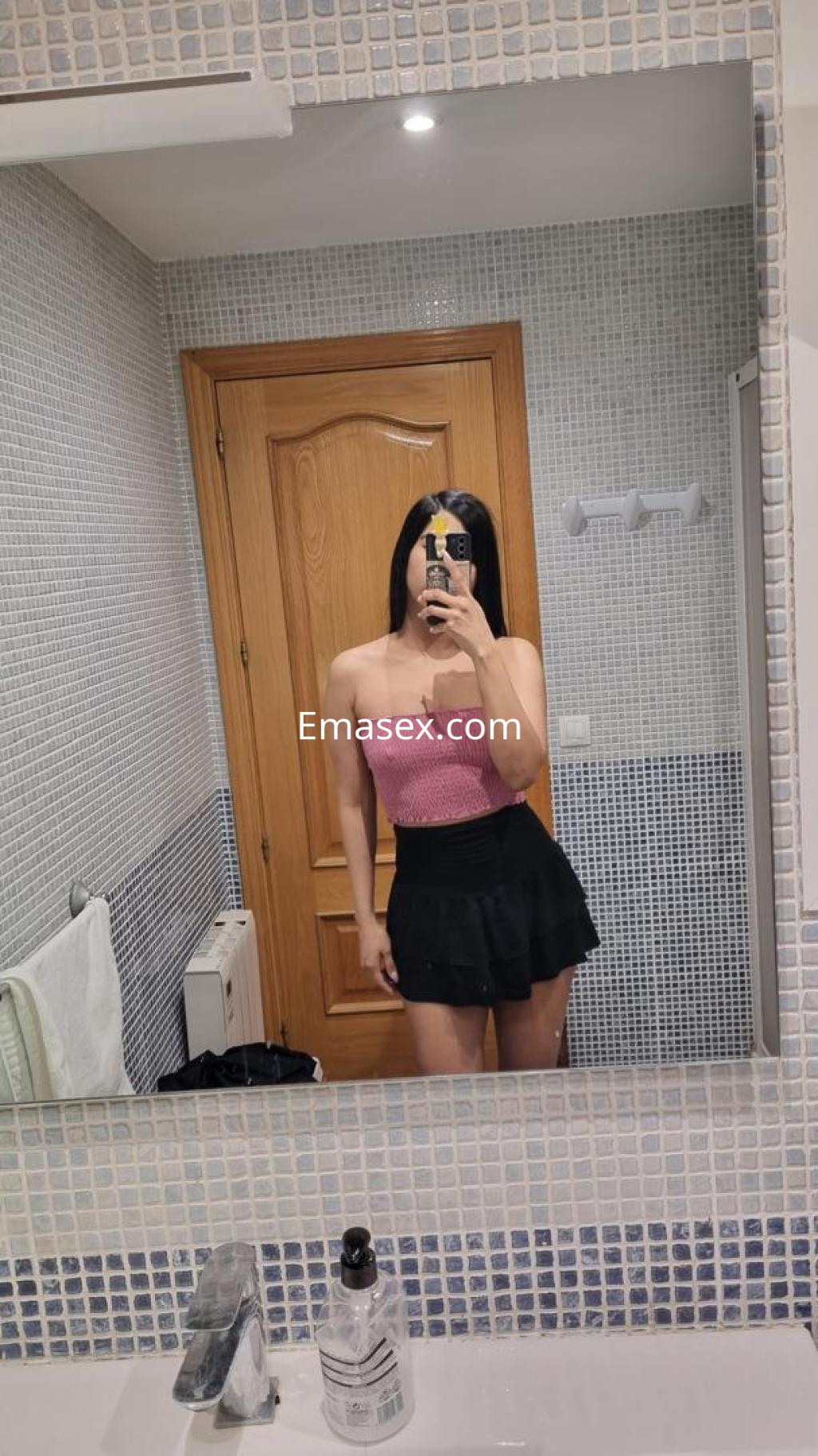 La puta se mira en el espejo, vistiendo un top rosa y una falda negra, en un baño moderno de Tarragona.