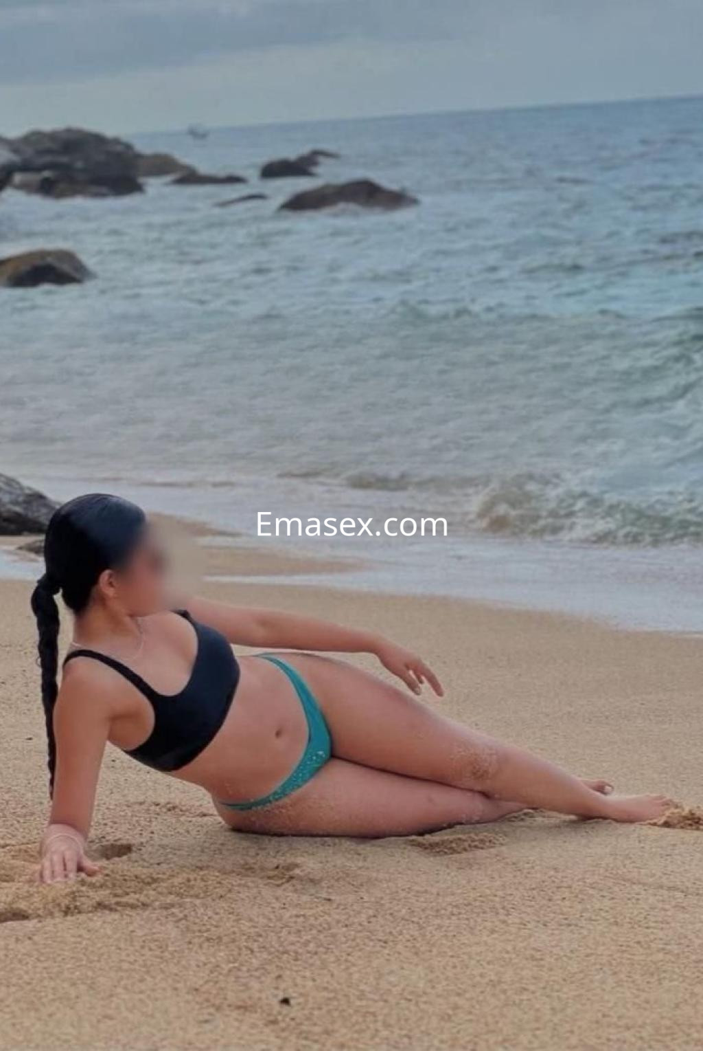 Puta en Lleida, recostada en la arena de la playa con bikini negro y verde, mirando hacia el mar.