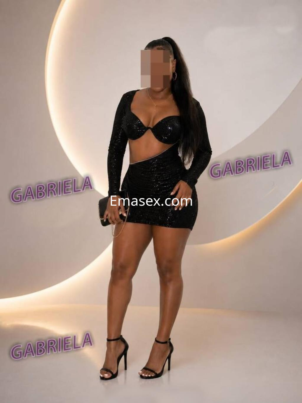 Puta en Valladolid con un vestido negro ajustado y escote, luciendo sandalias de tacón en un fondo iluminado.