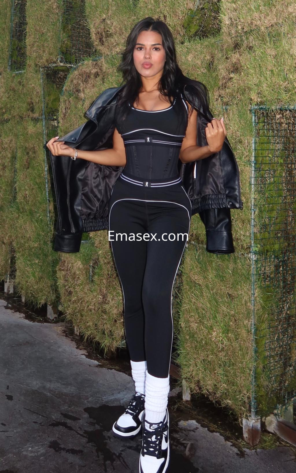 Escort en Vitoria viste un conjunto deportivo negro con detalles blancos, sosteniendo una chaqueta de cuero.