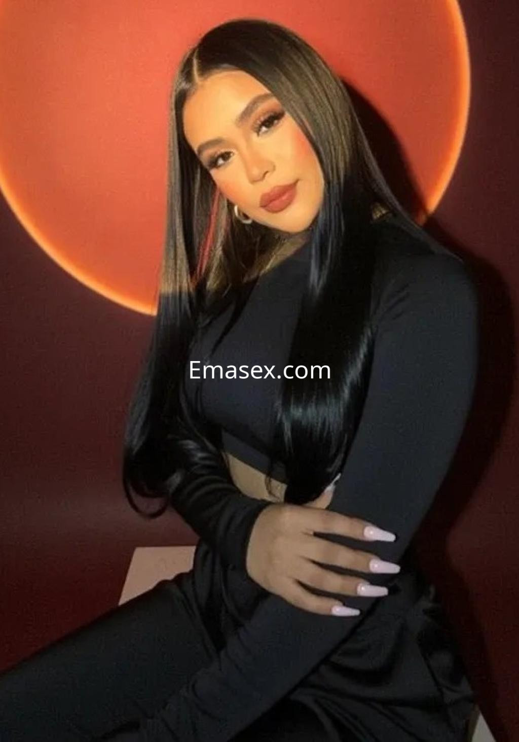 Escort en Coruña con cabello largo y lacio, vestida con un conjunto negro, posando con una mano en el brazo y fondo rojo.